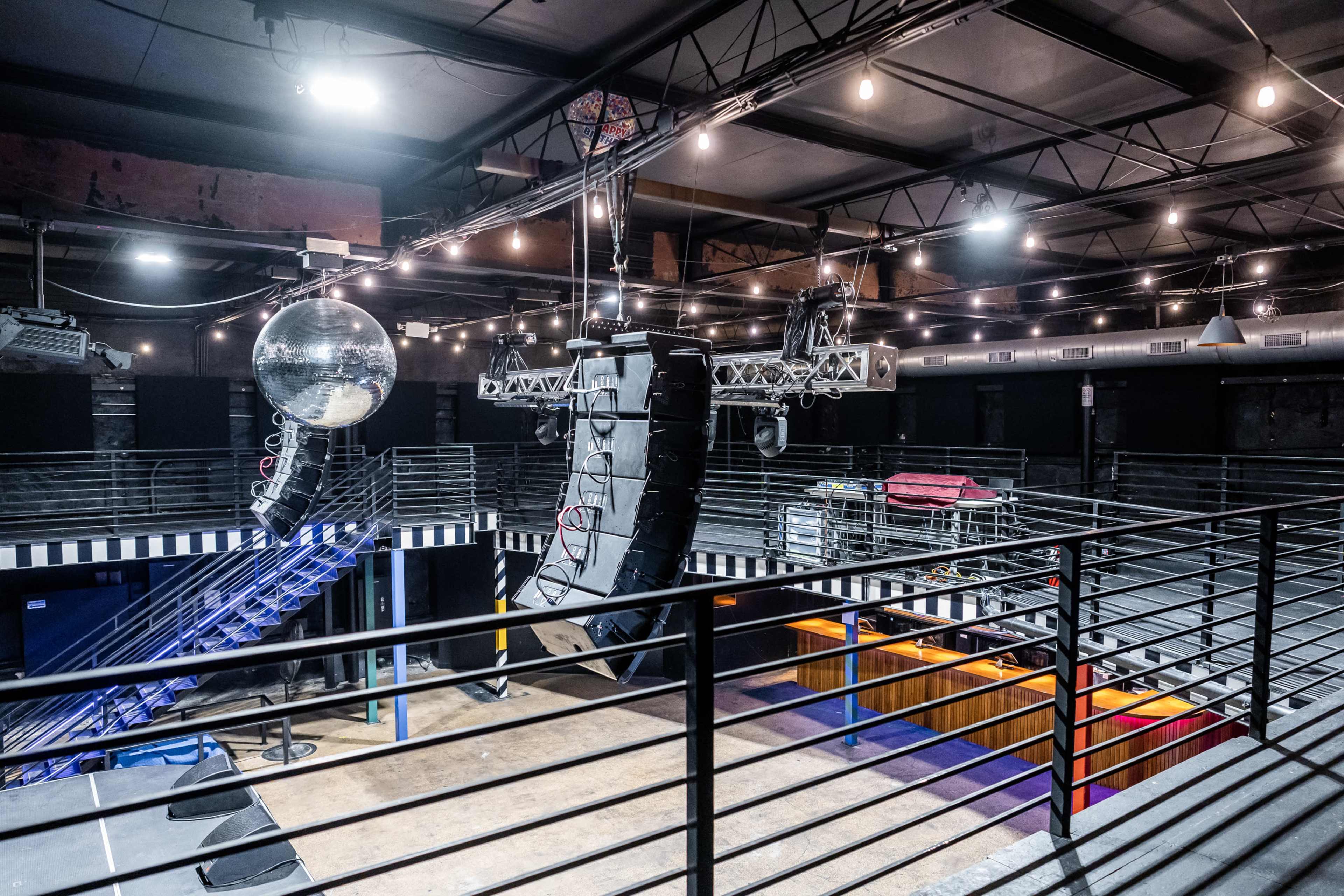An empty music venue with a stage, overhead sound equipment, and a disco ball reflecting light in the ceiling.