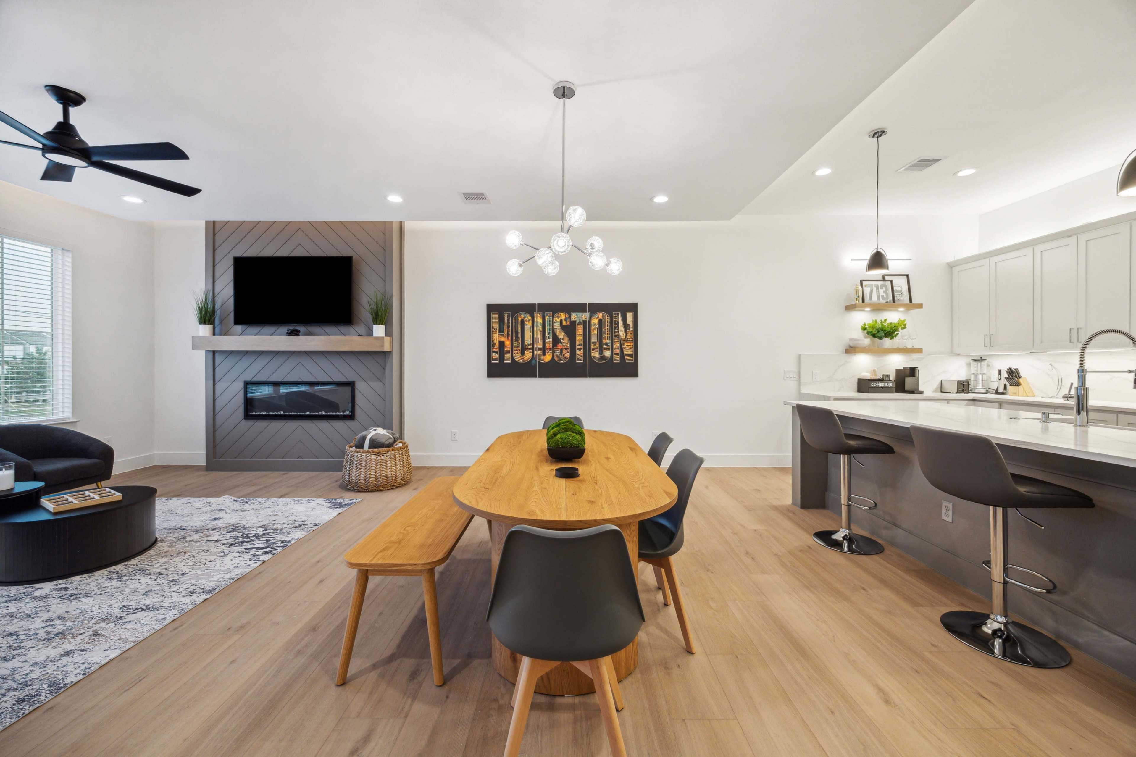 The image shows a modern living space with a dining table and chairs, a sectional couch, and a wall-mounted TV above a fireplace.