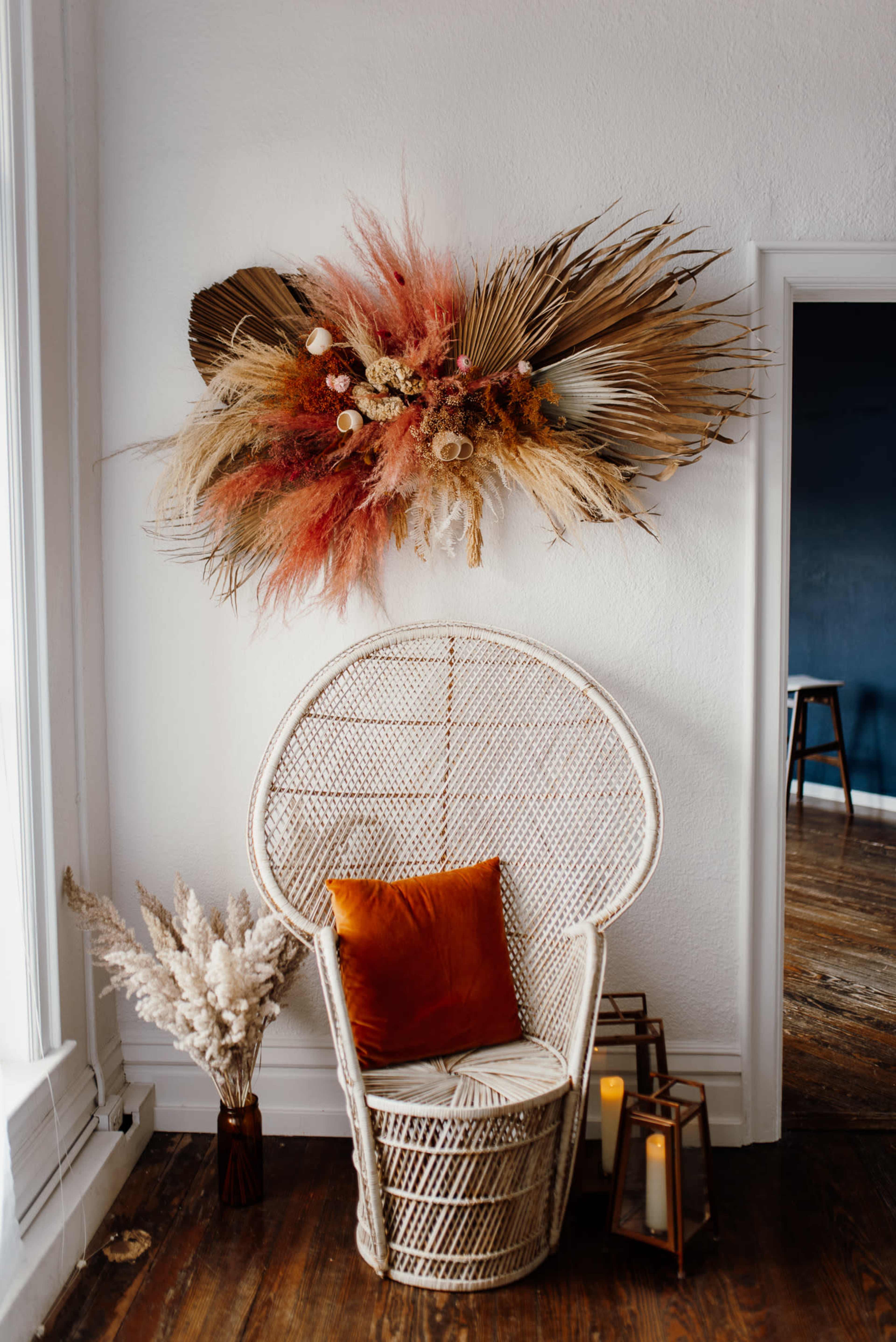 A rattan chair with an orange cushion is positioned next to a vase of dried flowers, beneath a decorative wall arrangement of pampas grass and preserved foliage.