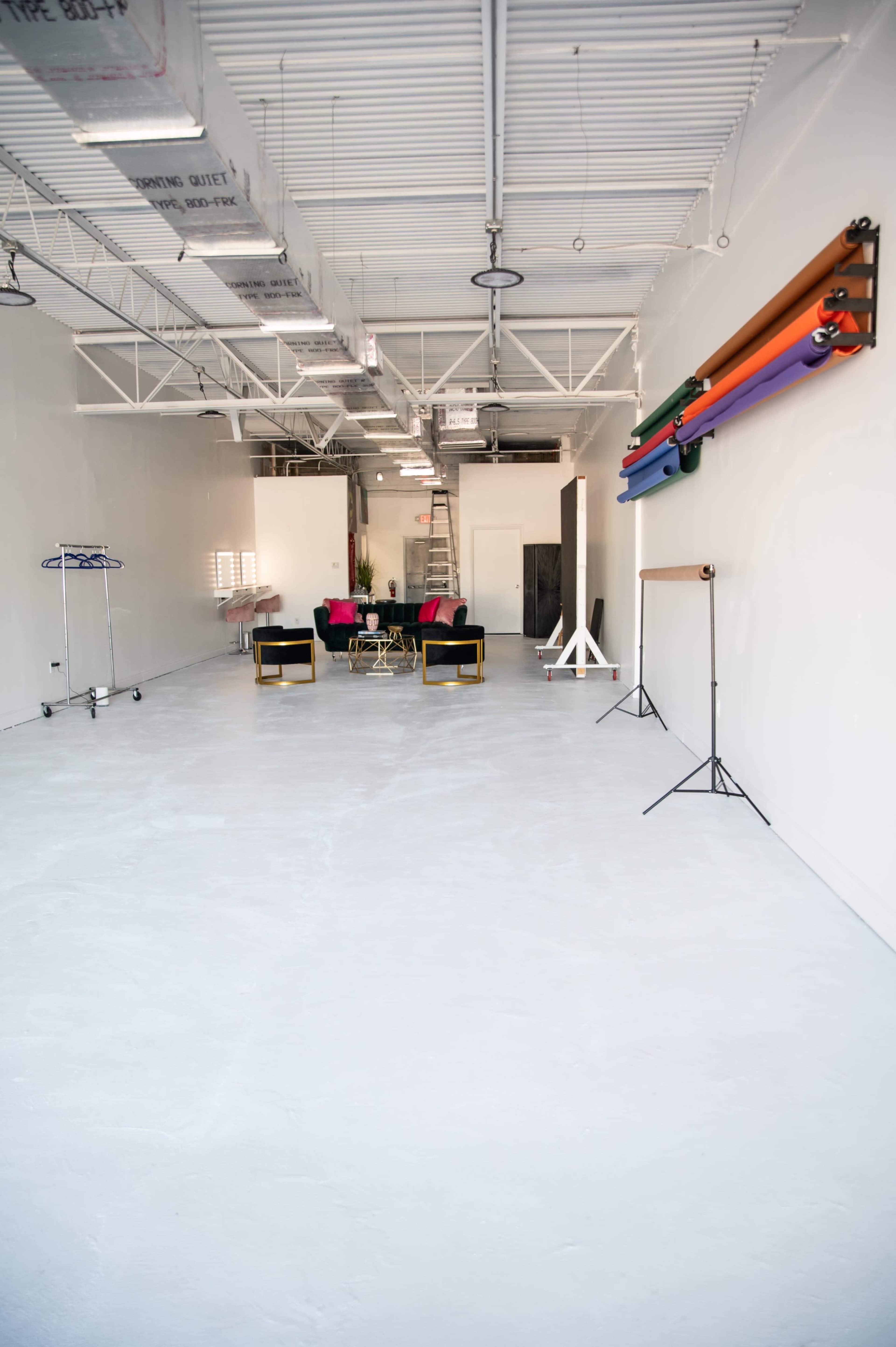 The image shows a spacious, empty room with a white floor and walls, featuring a seating area with chairs, a table, and several rolled-up fabric displays on a rack against one wall.