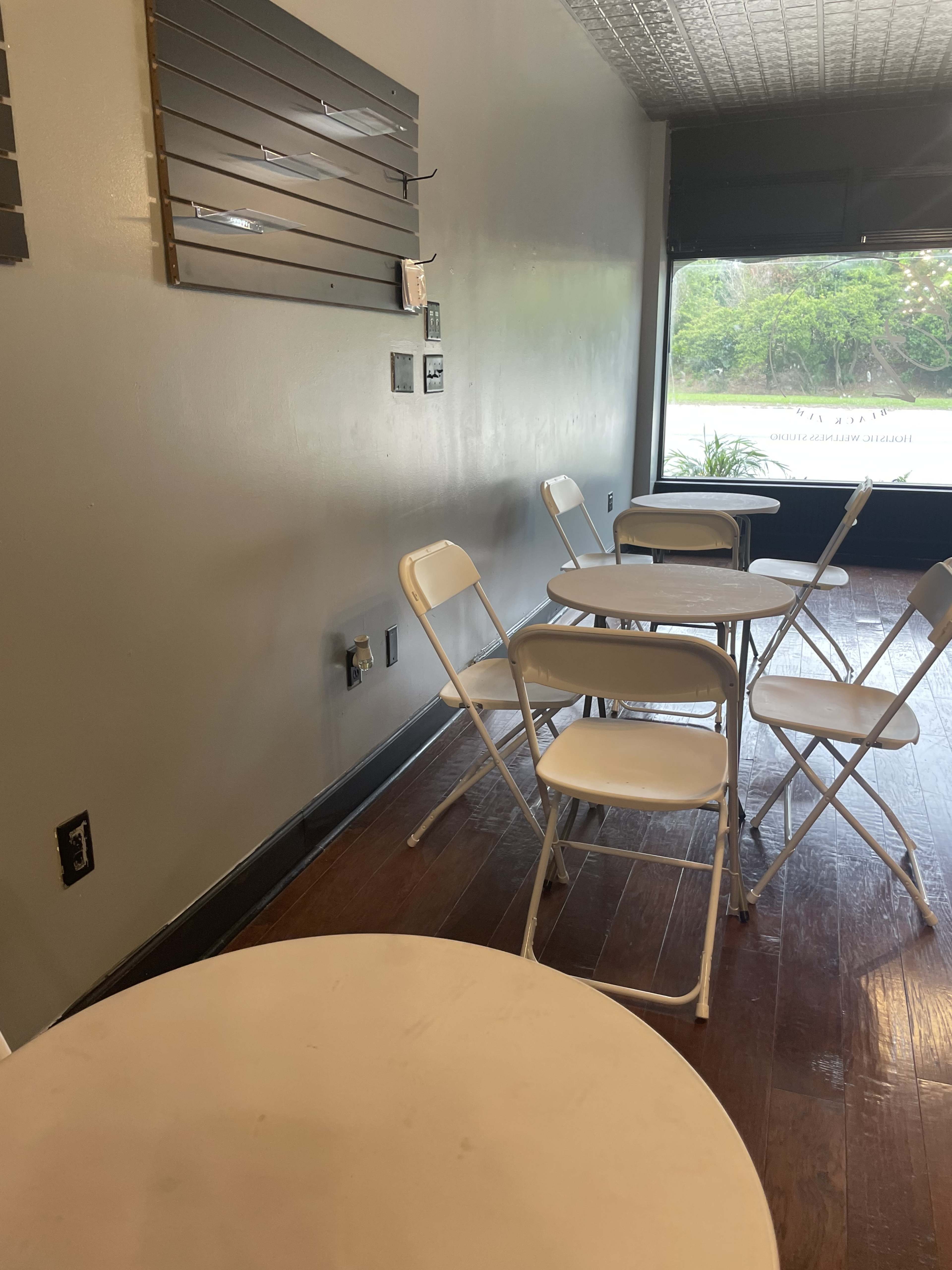 The image shows a small, sparsely furnished interior with white folding tables and chairs arranged on a wooden floor.