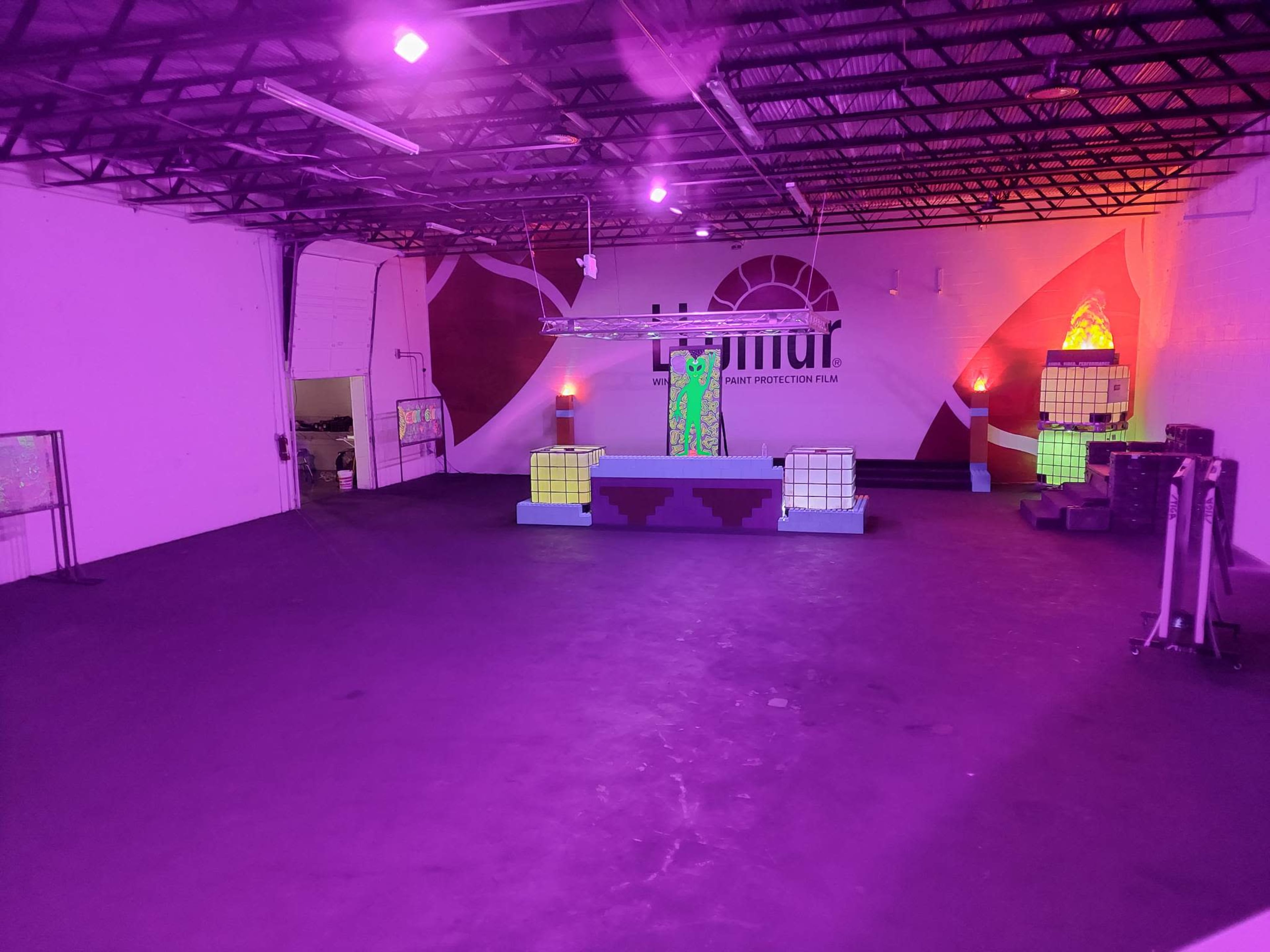 An empty indoor space with purple lighting, featuring illuminated geometric structures and a stage area against a mural on the wall.