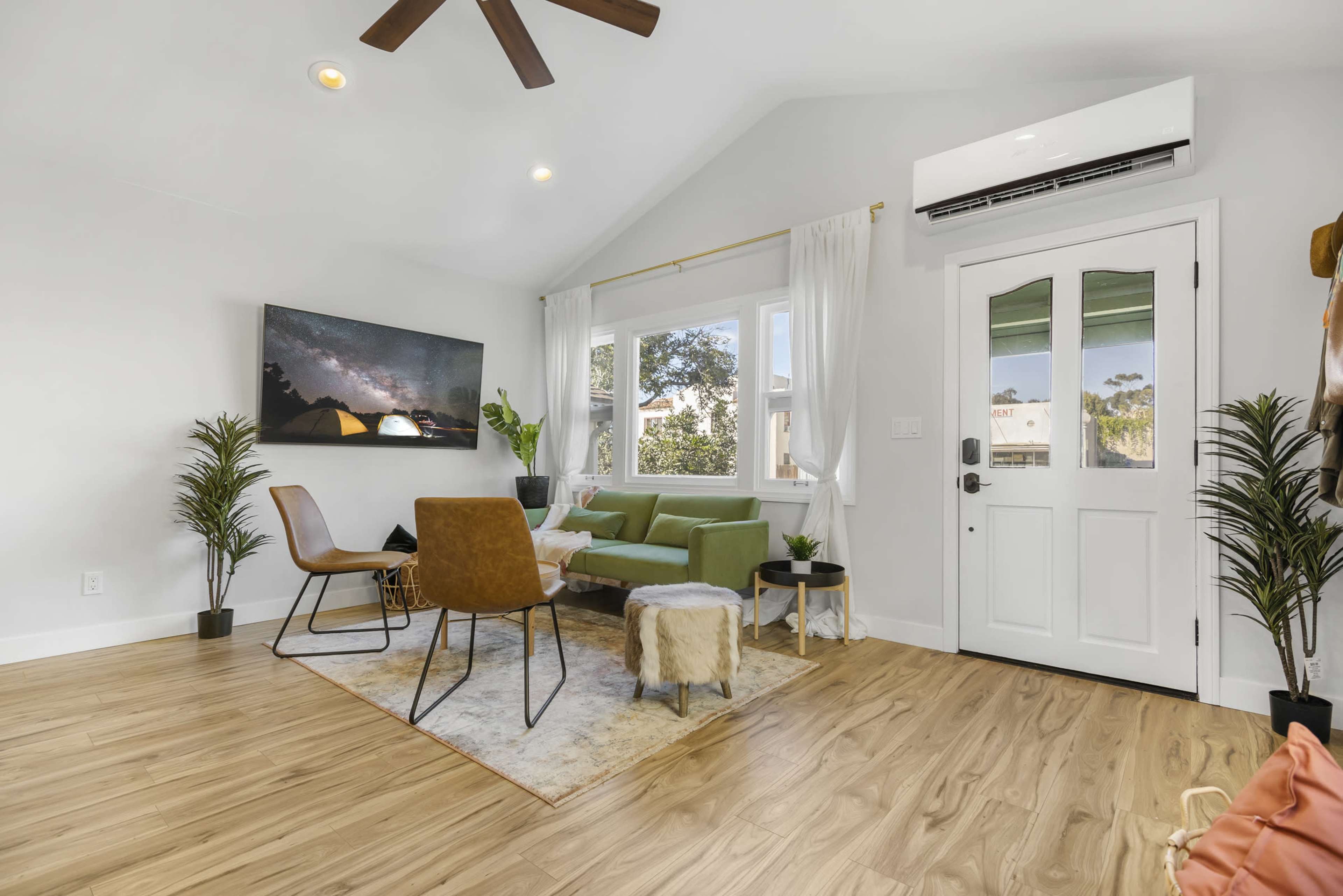 The room features a green sofa, a wooden floor, a wall-mounted TV, and large windows, creating a bright and open living area.