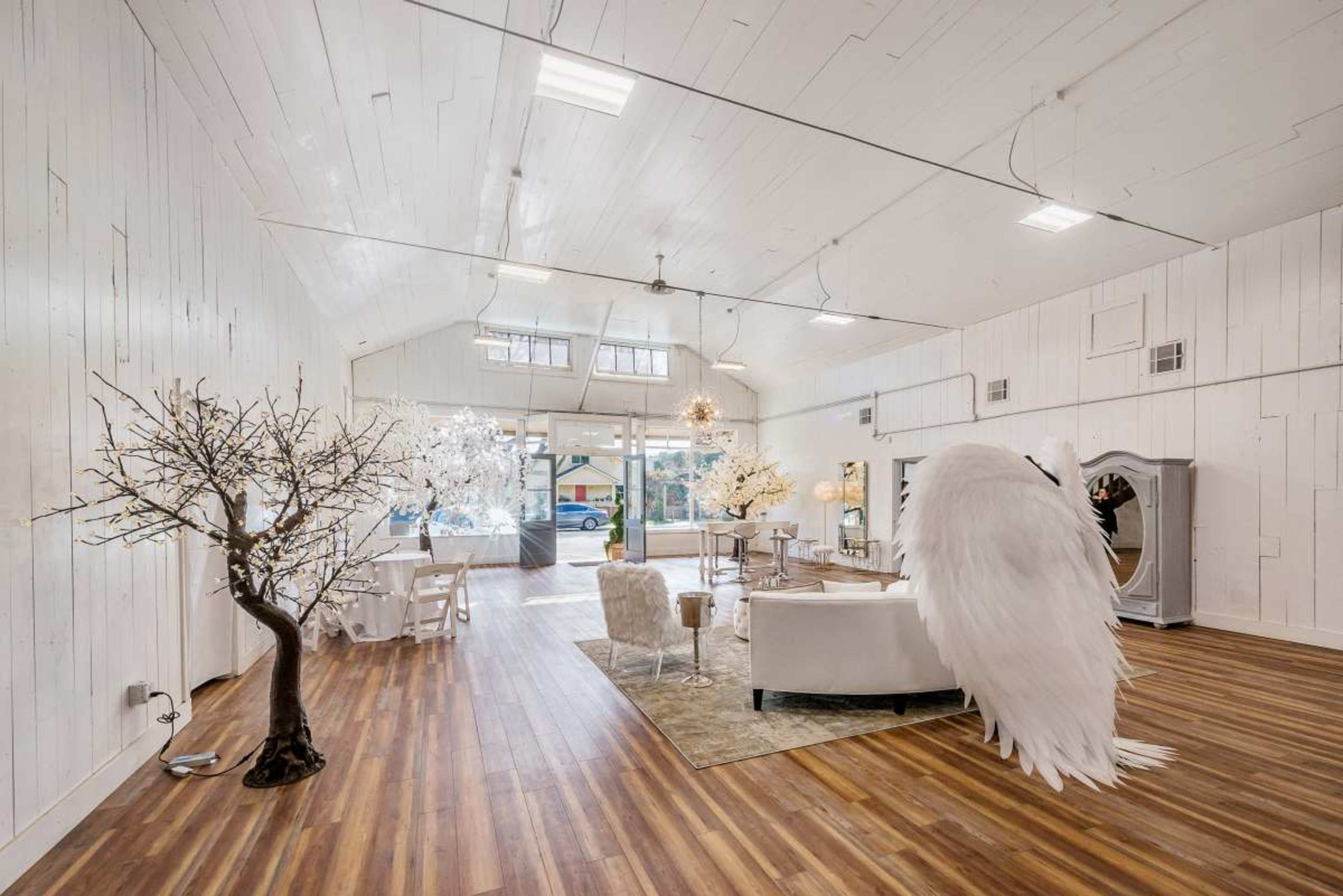 The image shows a bright, airy interior space with white walls, wooden flooring, and various decorative elements, including tree sculptures and a large white winged installation.