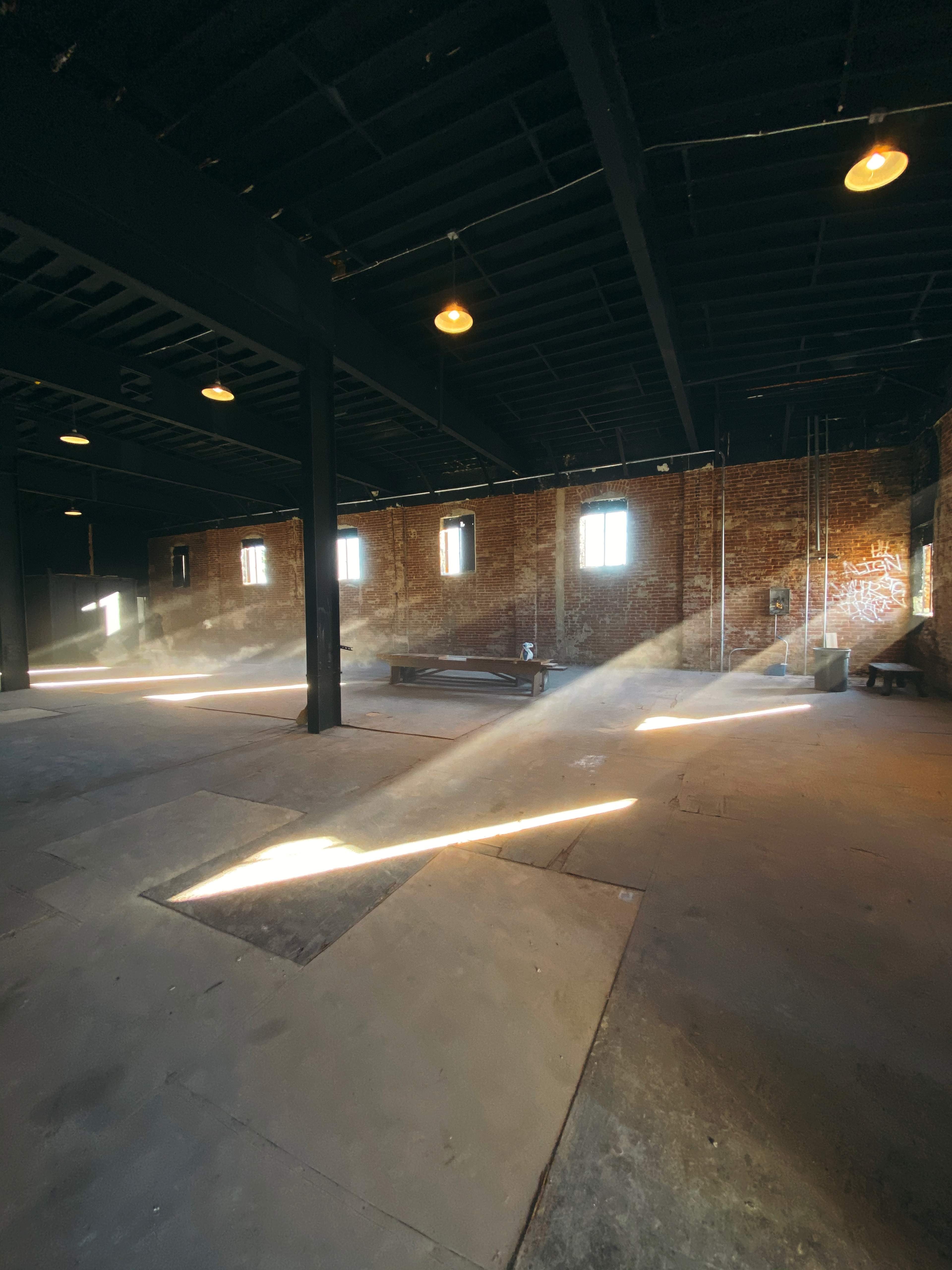 Sunlight streams through the windows of a spacious industrial room, illuminating dust particles in the air and highlighting the brick walls.