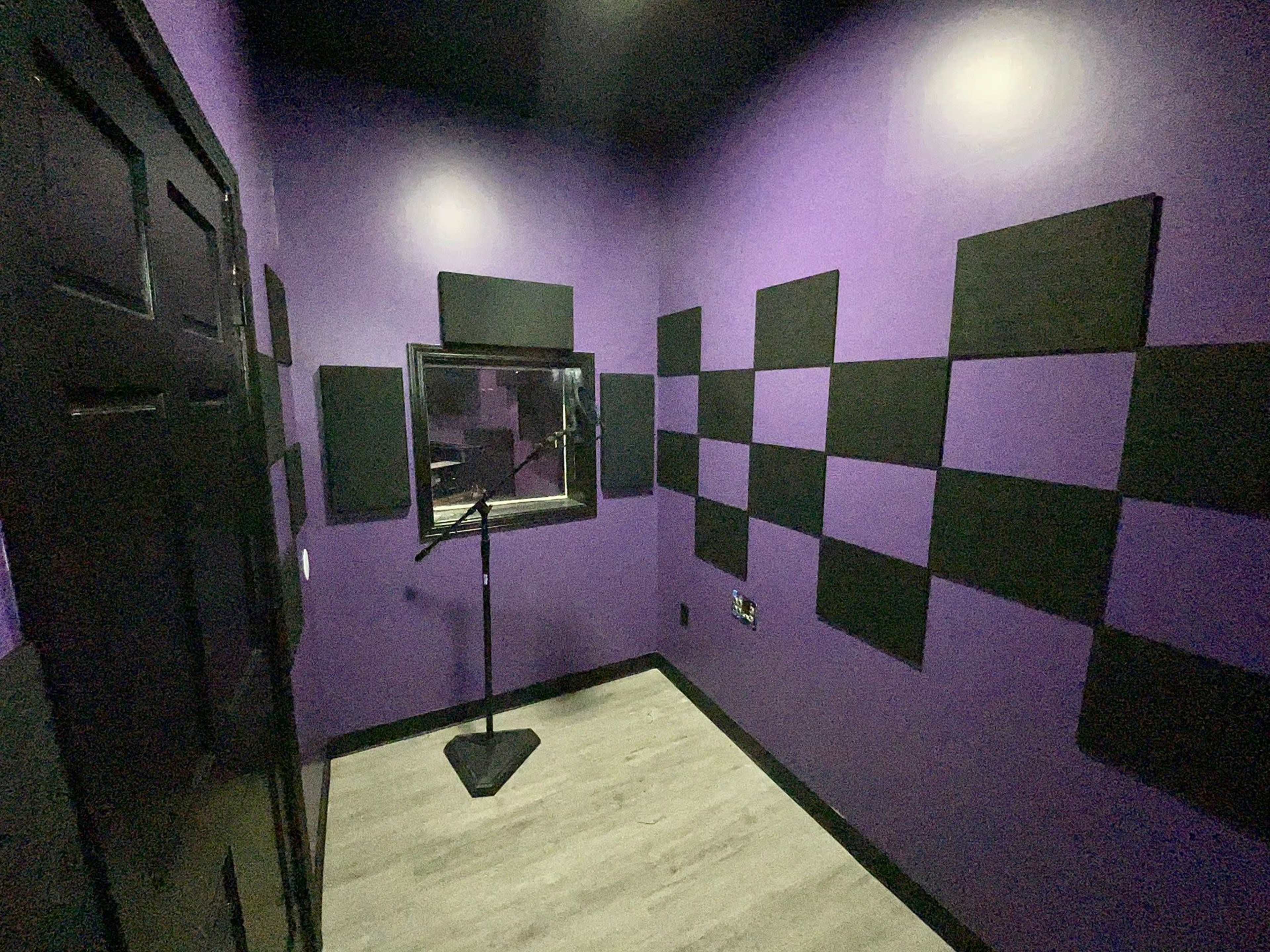 The image shows a small recording studio with purple walls, black acoustic panels, a microphone stand, and a window to an adjacent room.