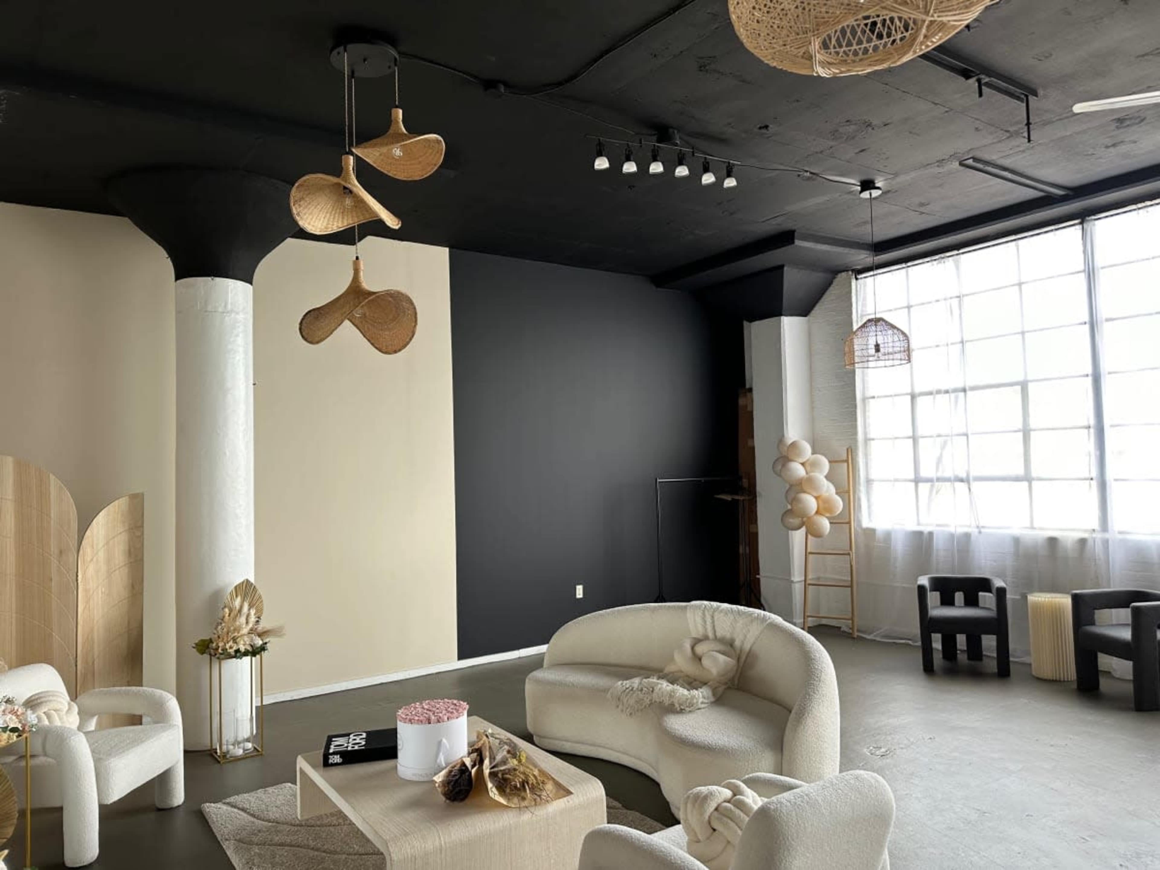 The image shows a modern interior space with beige and dark walls, featuring circular seating arrangements, decorative elements, and large windows allowing natural light.