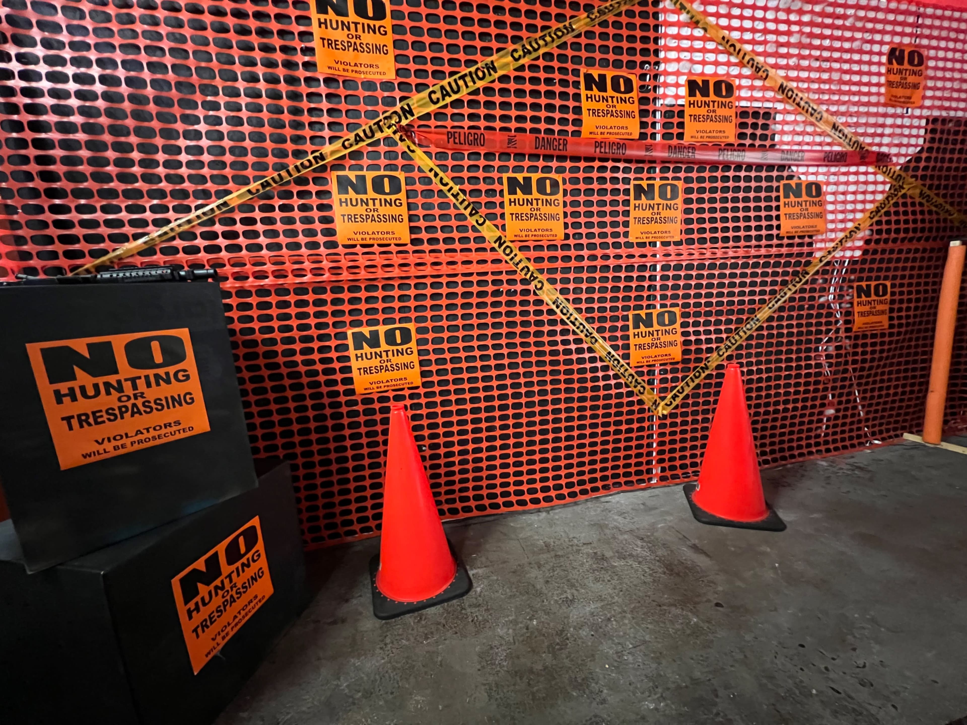 The image shows a wall covered in bright orange "No Hunting" and "No Trespassing" signs, flanked by two red traffic cones and a section of caution tape.