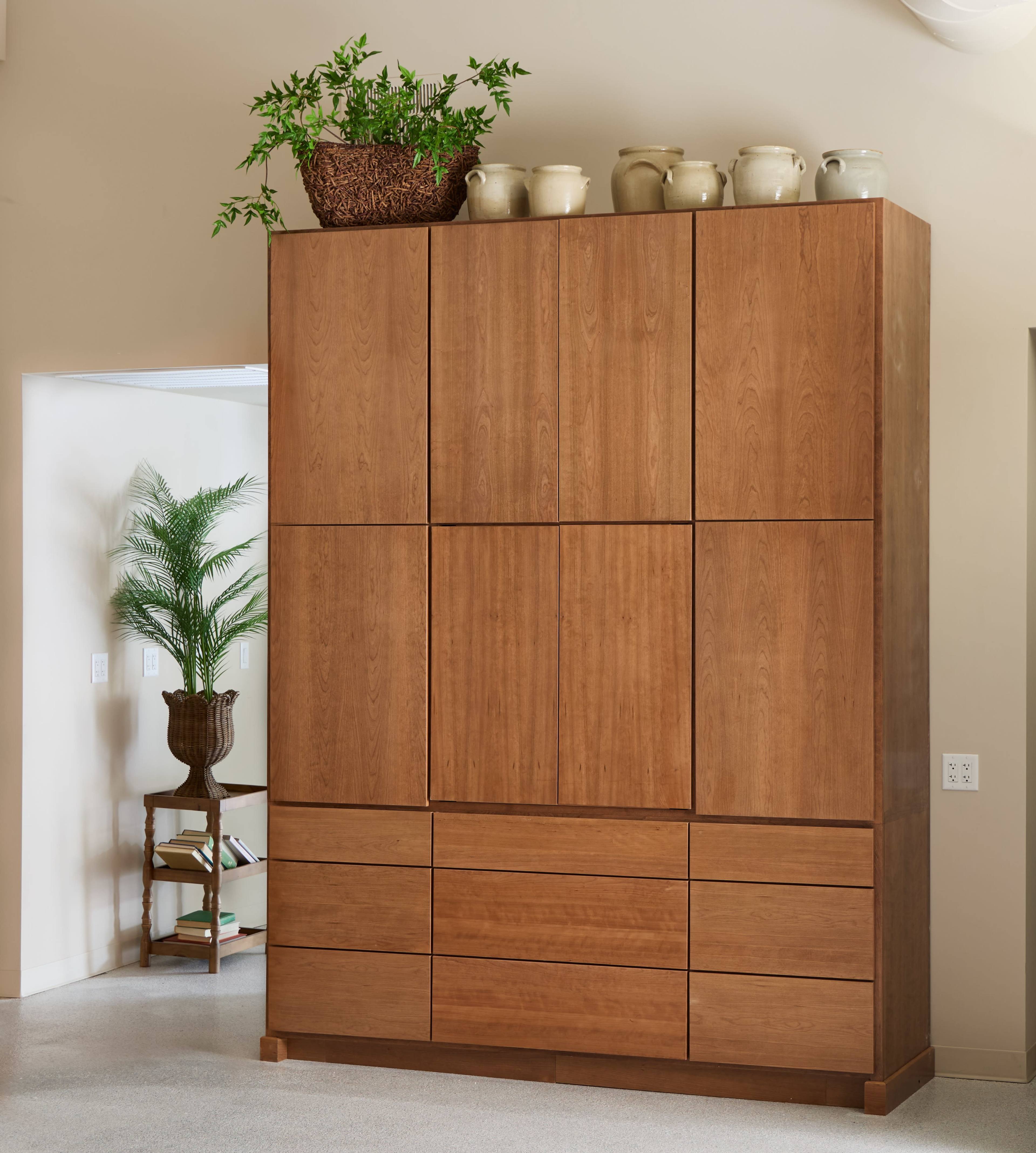 The image shows a tall wooden cabinet with a smooth finish, topped with a planter and several ceramic pots, set in a modern interior space.