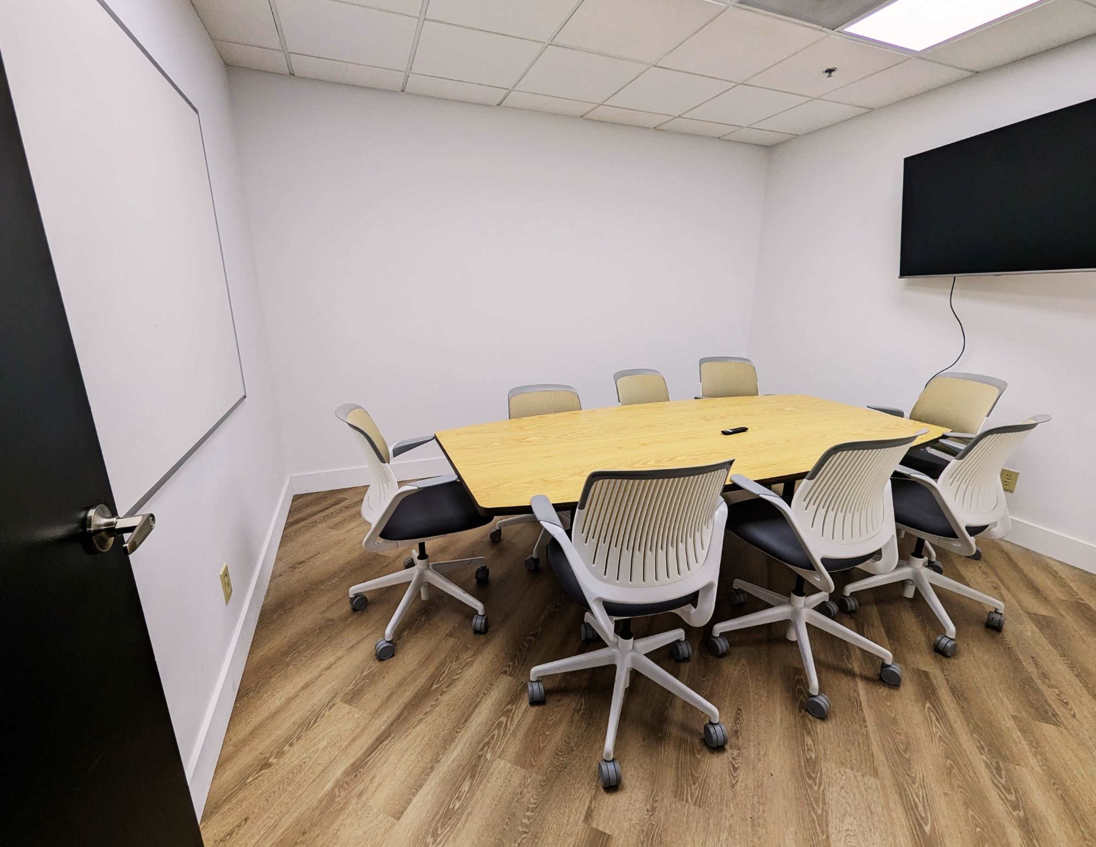 A conference room features a large wooden table surrounded by eight white chairs, with a mounted screen and a whiteboard on the walls.