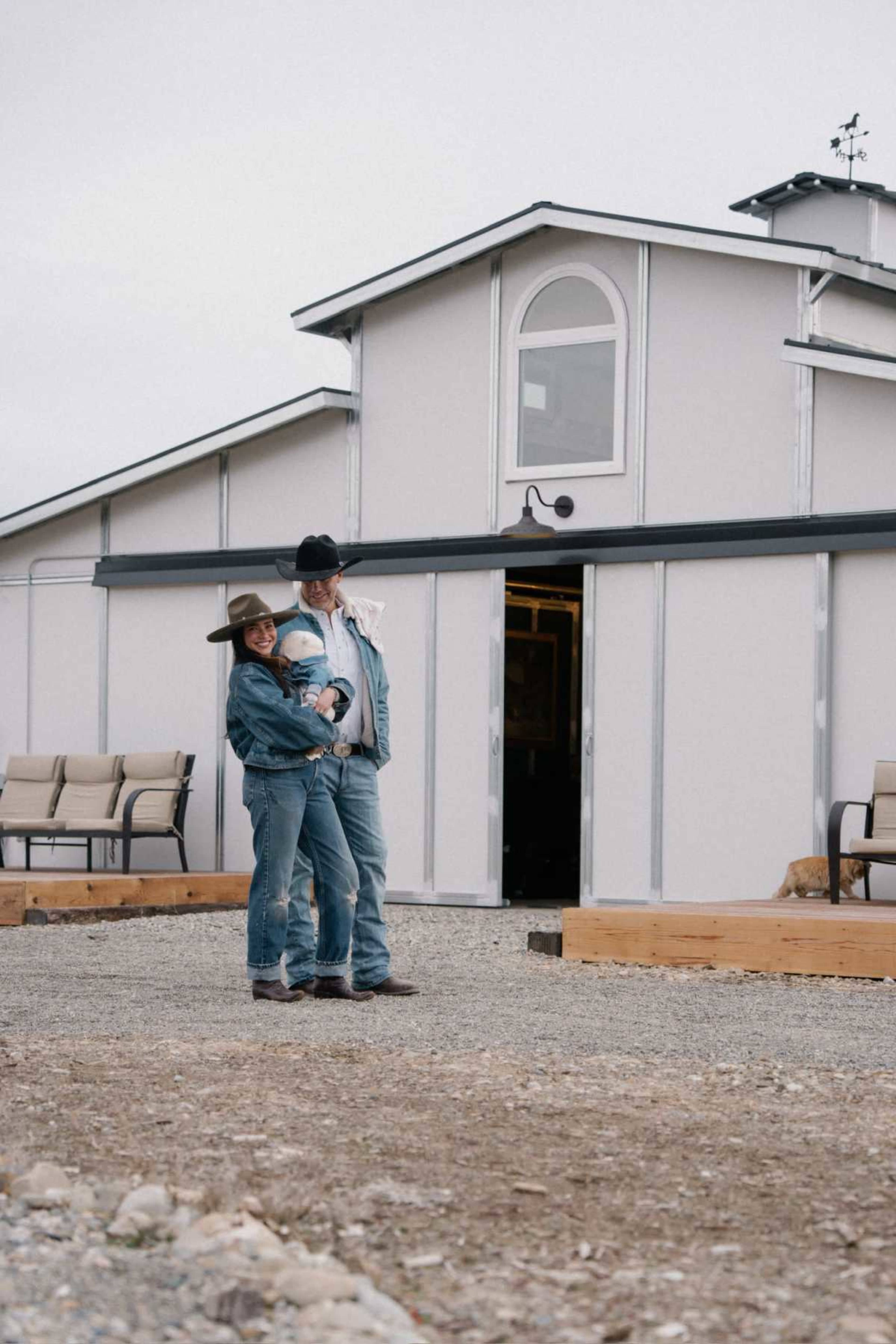 A man and woman stand outside a building, holding a child, with seating arranged on the gravel in front of them.