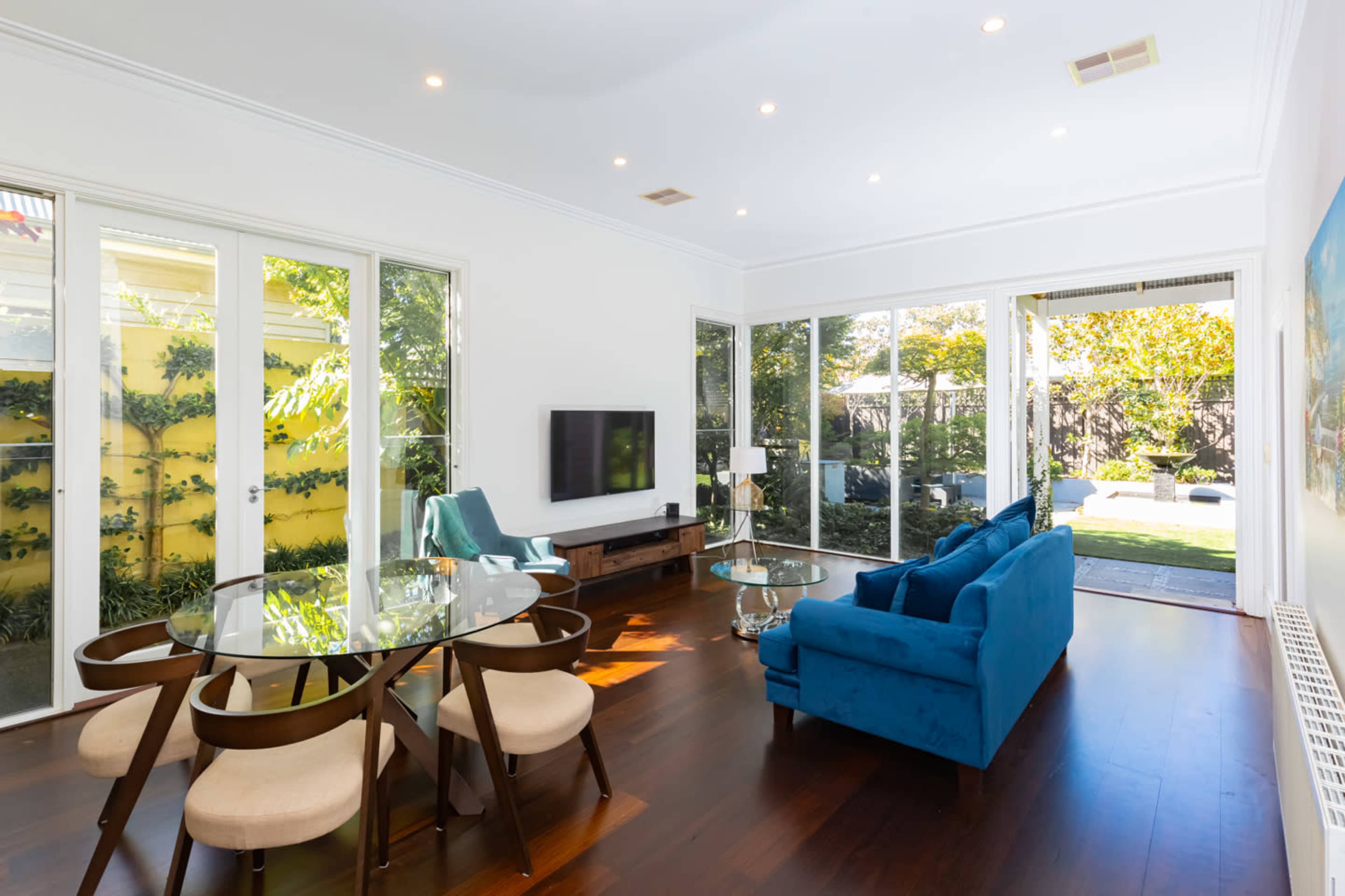 A modern living room features a glass dining table, a blue sofa, and large windows displaying a garden view.