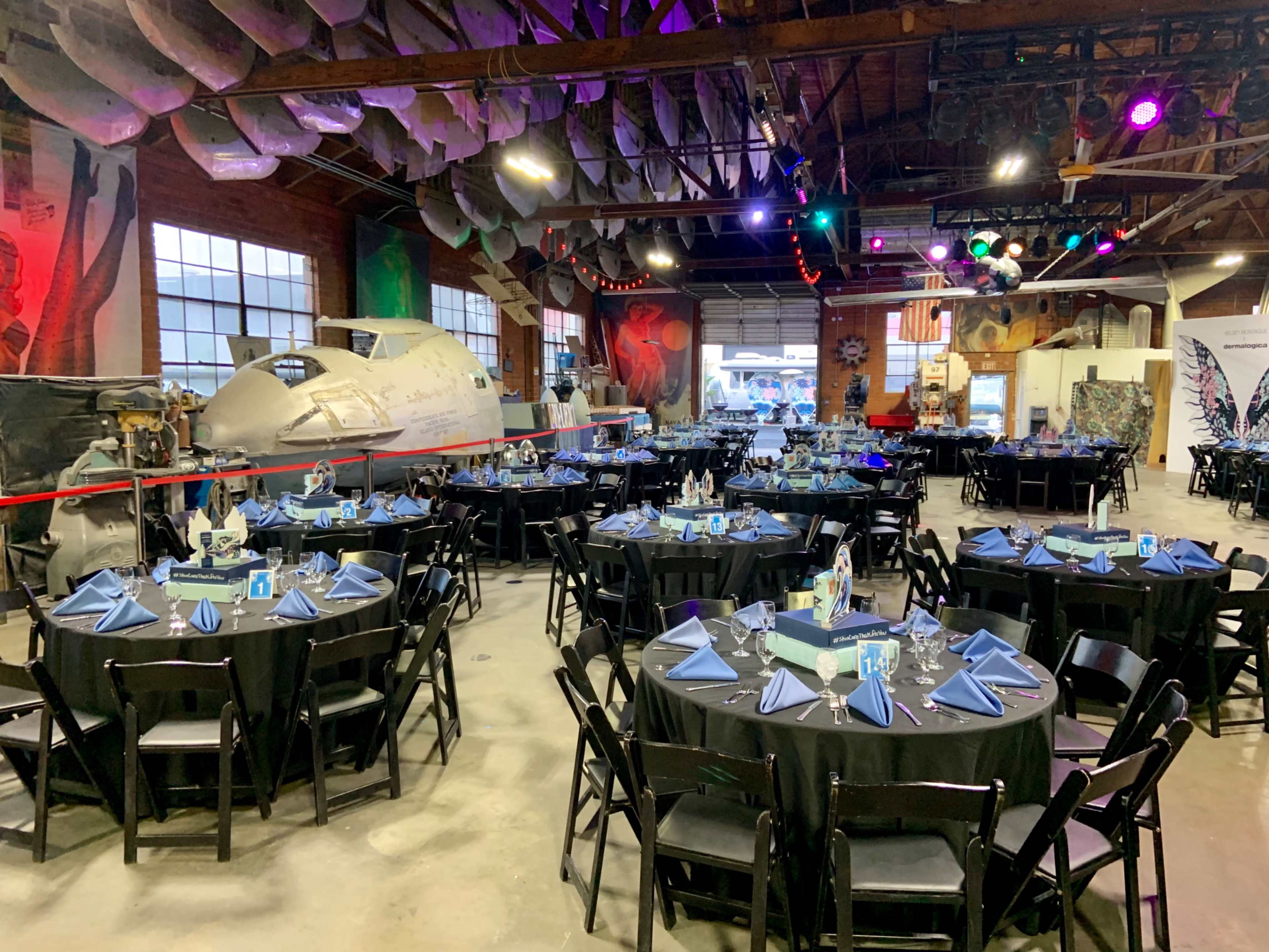 The image shows a large industrial space set up with multiple round tables covered in black tablecloths, each decorated for an event, alongside an aircraft and colorful lighting in the background.
