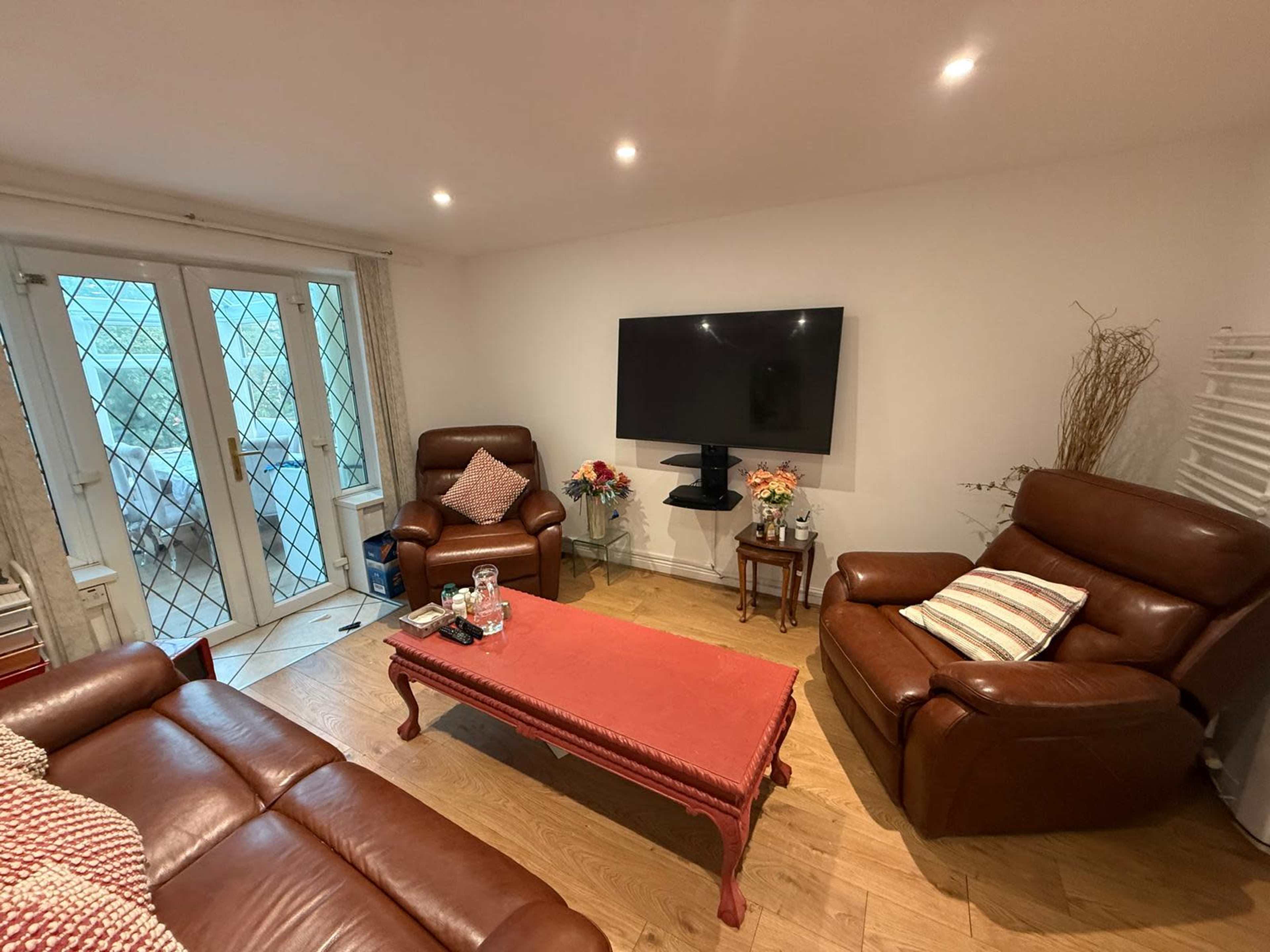 The image shows a living room with two brown leather chairs, a red coffee table, a flat-screen TV mounted on the wall, and large glass doors leading to an outdoor area.