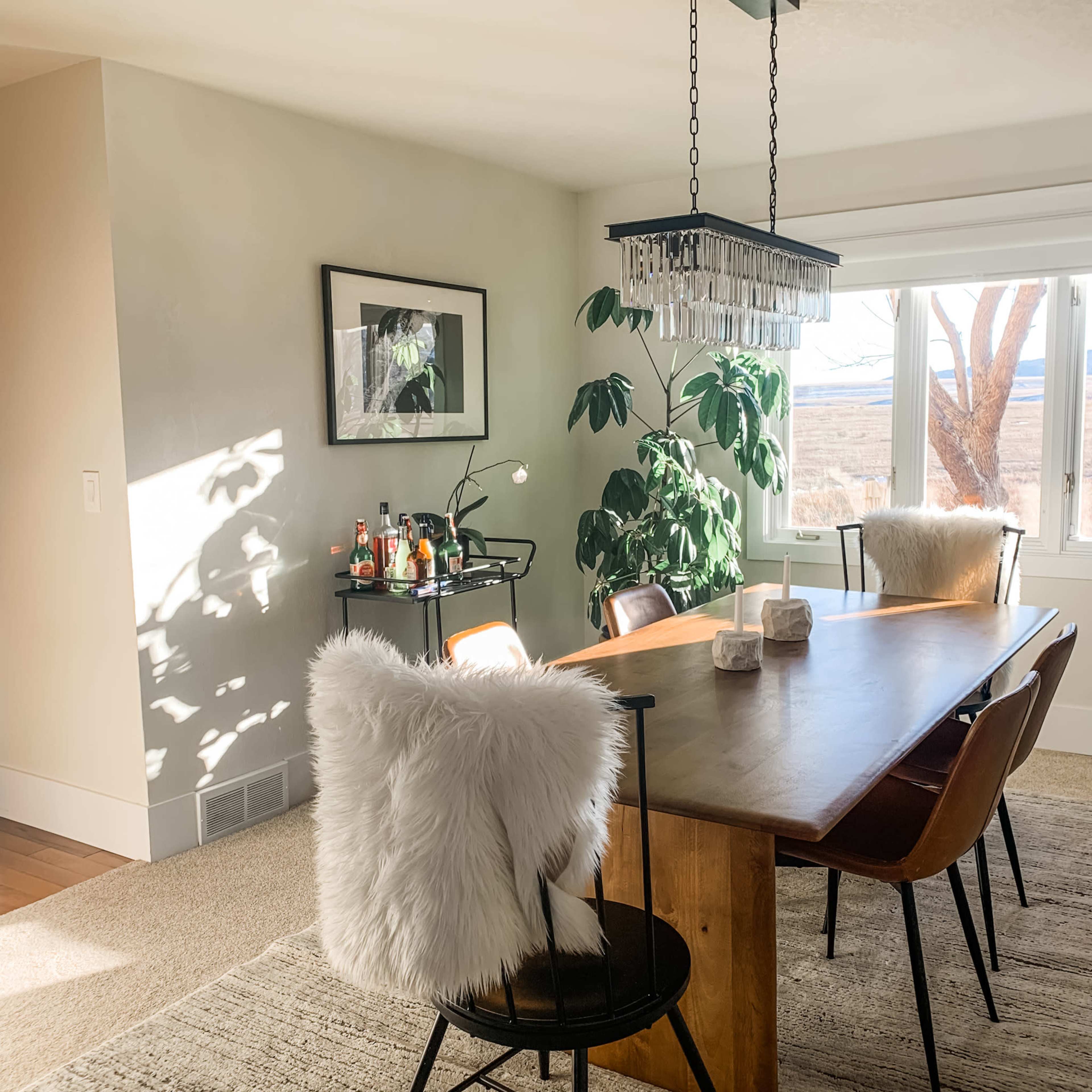 A dining area features a wooden table surrounded by chairs, with a plant and bar cart visible near a window.