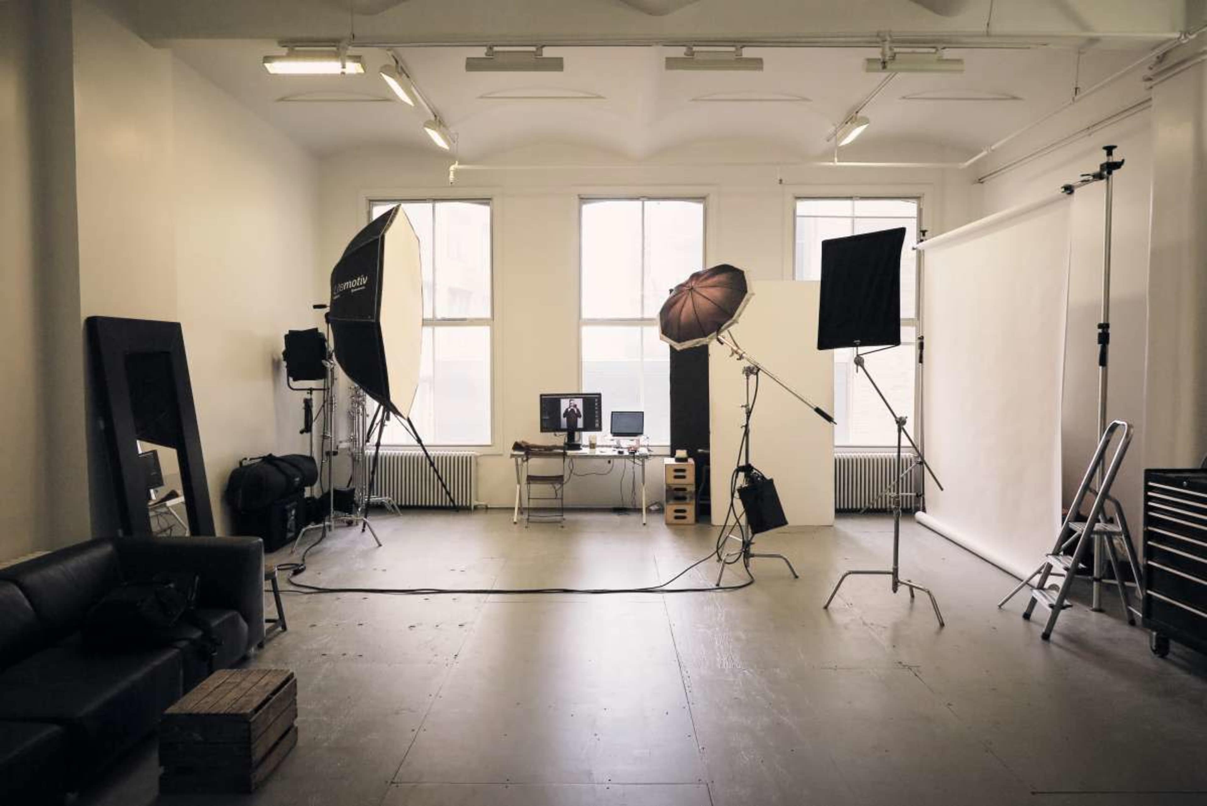 A spacious photography studio features lighting equipment, a desk with a computer, and large windows illuminating the interior.
