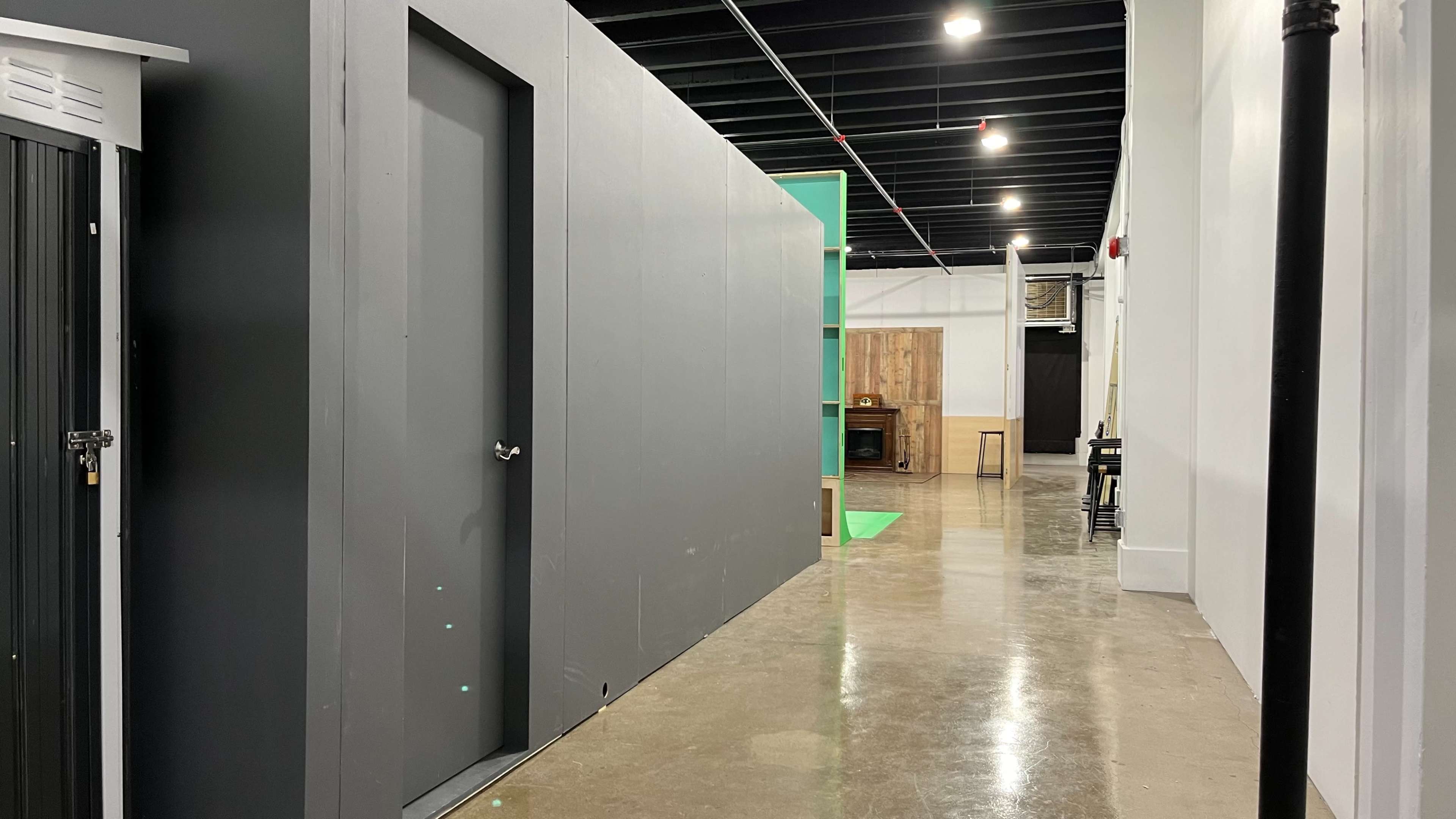 The image shows a narrow hallway featuring a gray wall, a closed door on the left, and visible green and wooden elements at the far end.