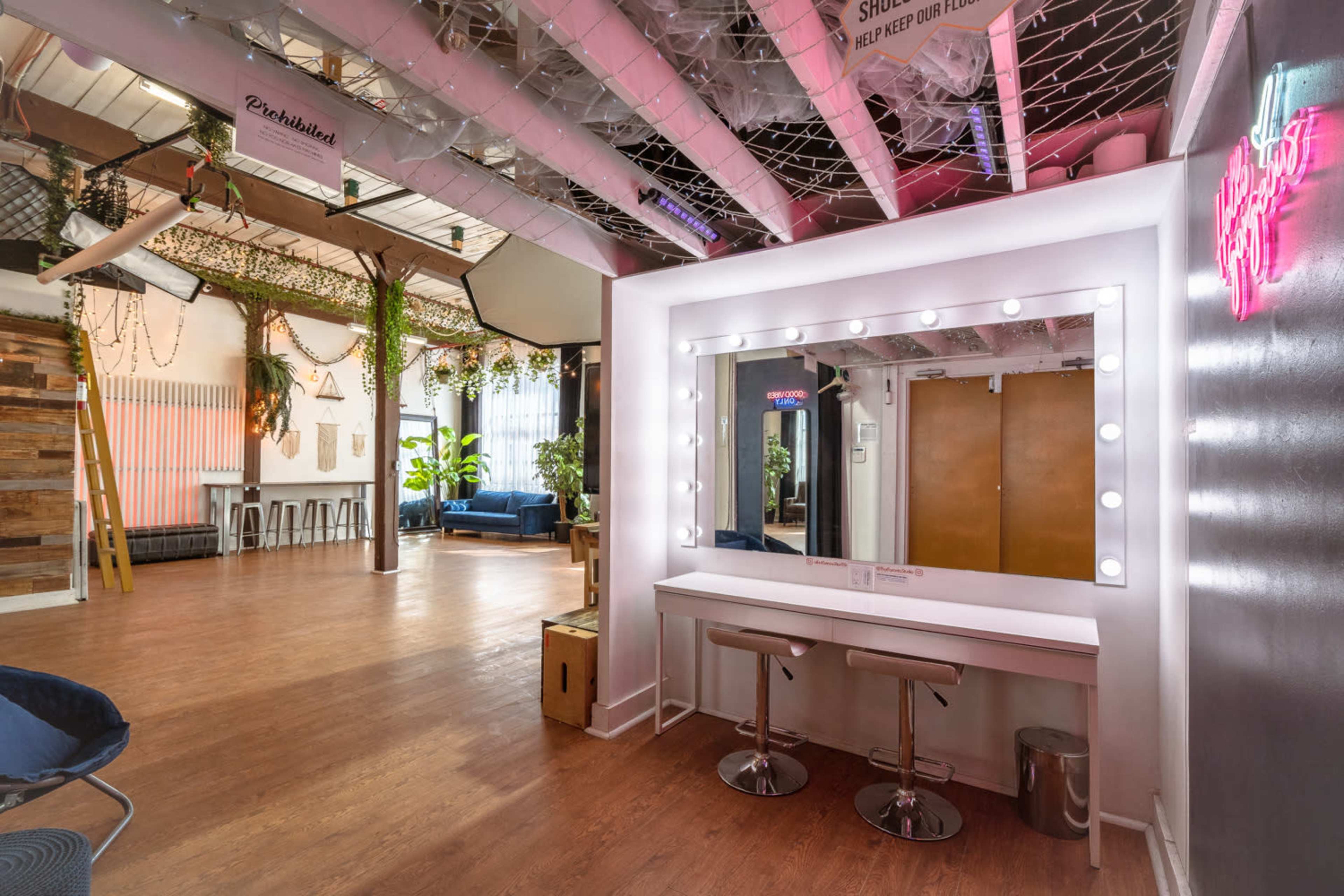 A bright, open space featuring a vanity mirror with lights, surrounded by decorative plants and modern furniture, with a wood-paneled wall and a seating area in the background.