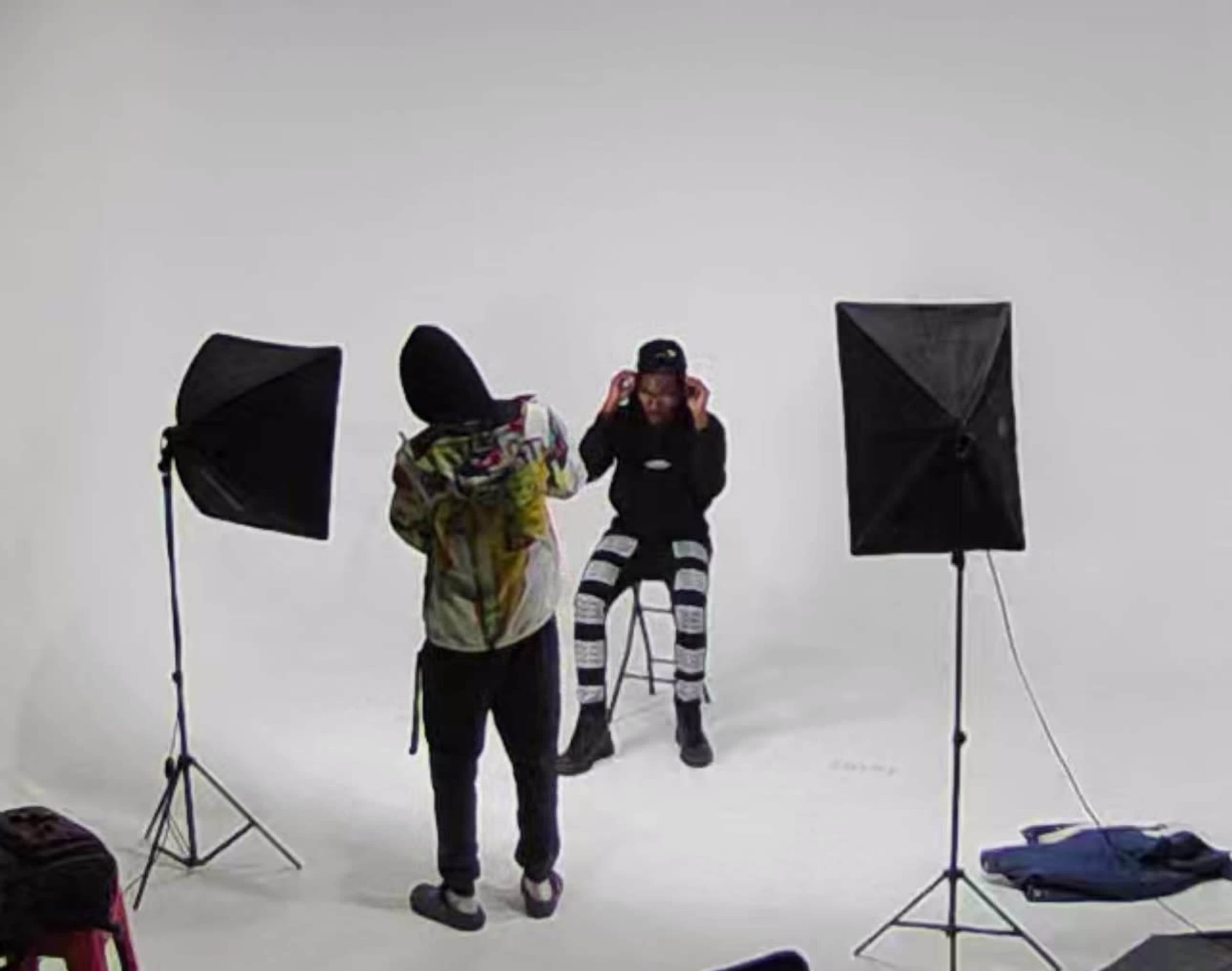 A person wearing a hoodie prepares to photograph another person seated on a stool, with studio lighting set up around them.