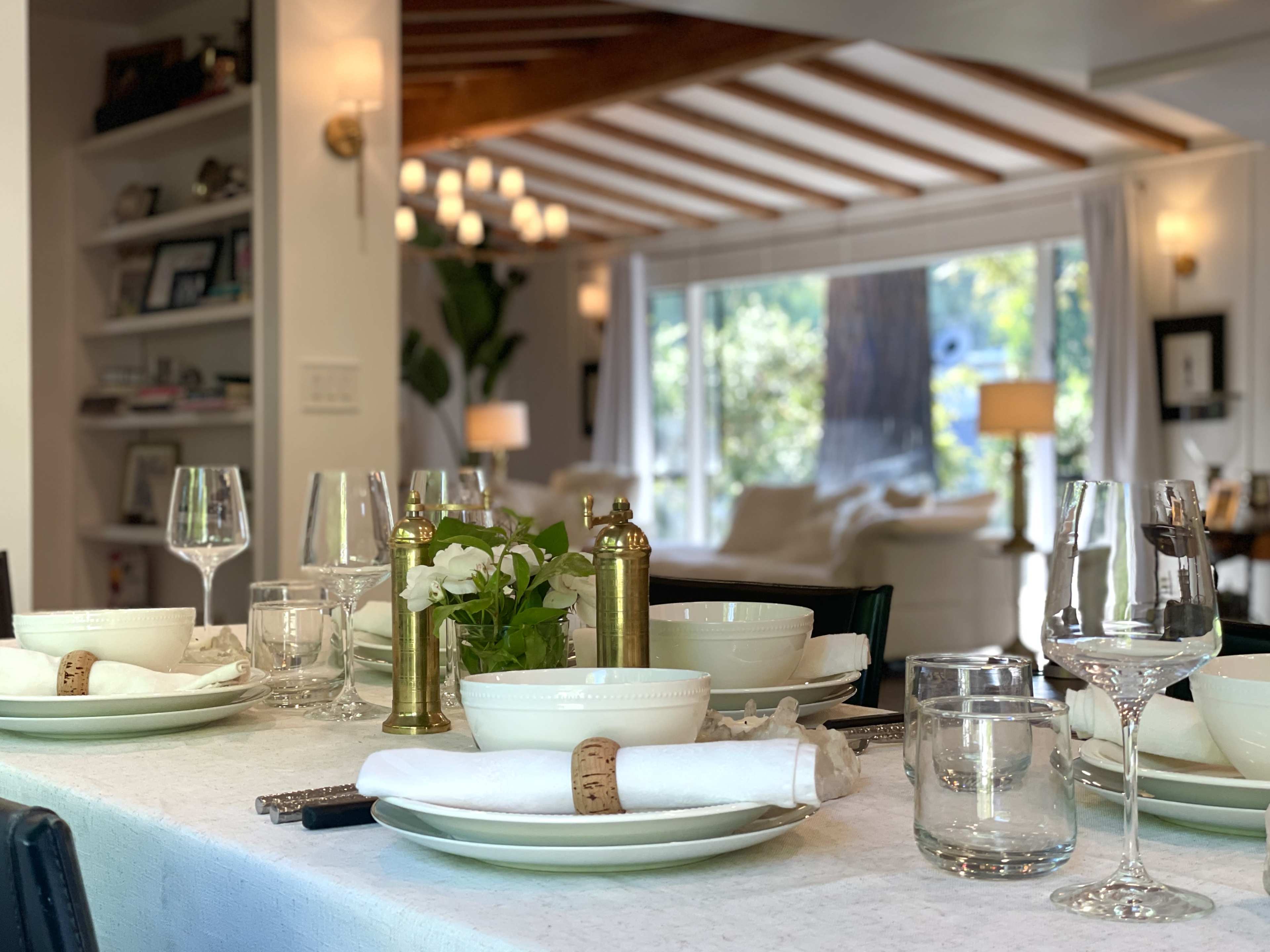 A dining table is set with elegant dishware, glassware, and a small arrangement of greenery, with a bright living area visible in the background.