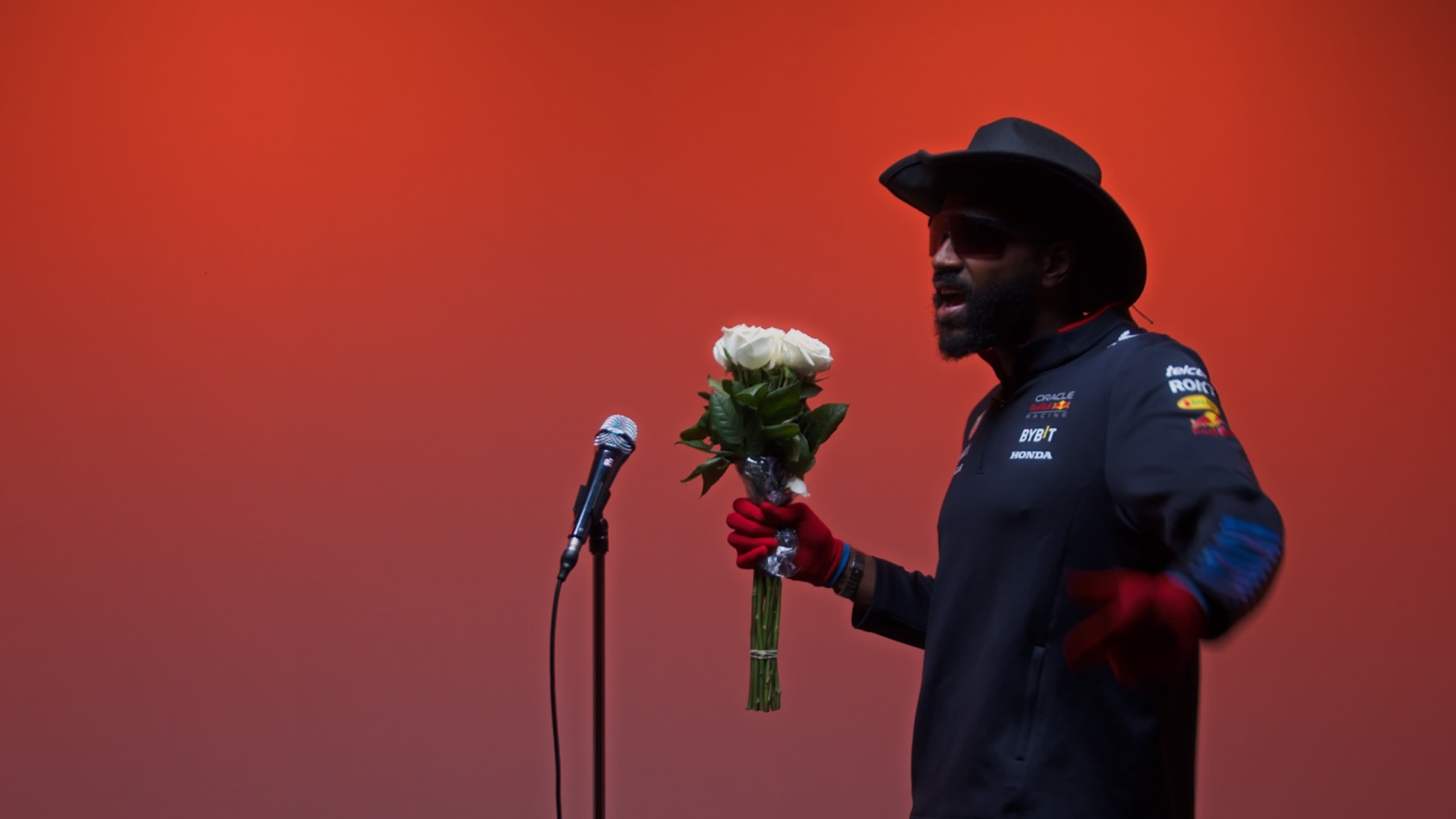 A man wearing a cowboy hat and red gloves holds a bouquet of white roses while standing at a microphone against a warm orange background.