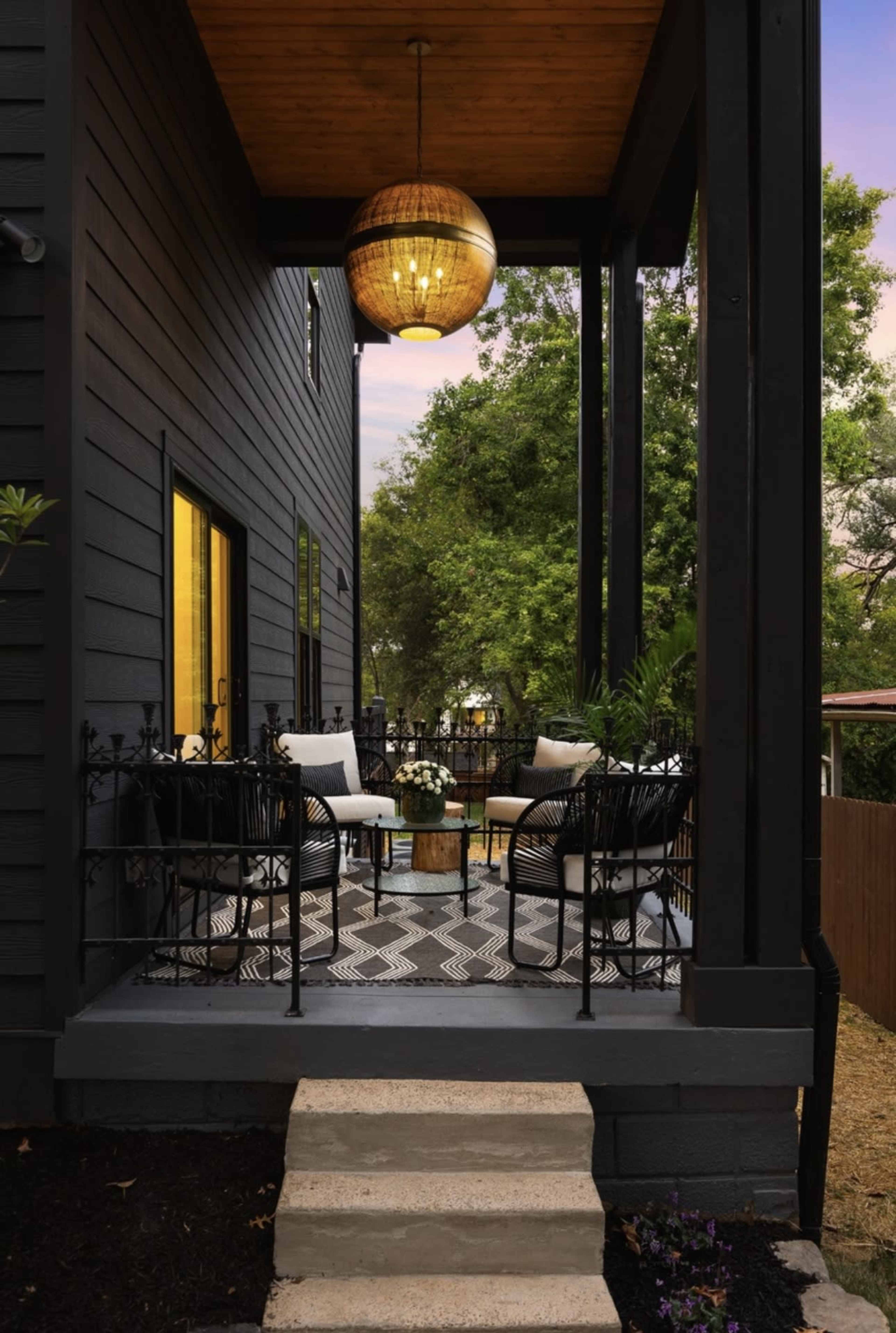 The image shows a front porch area with a patterned tile floor, two black chairs, and a round table, surrounded by a black wrought-iron railing and lit by a hanging pendant light.