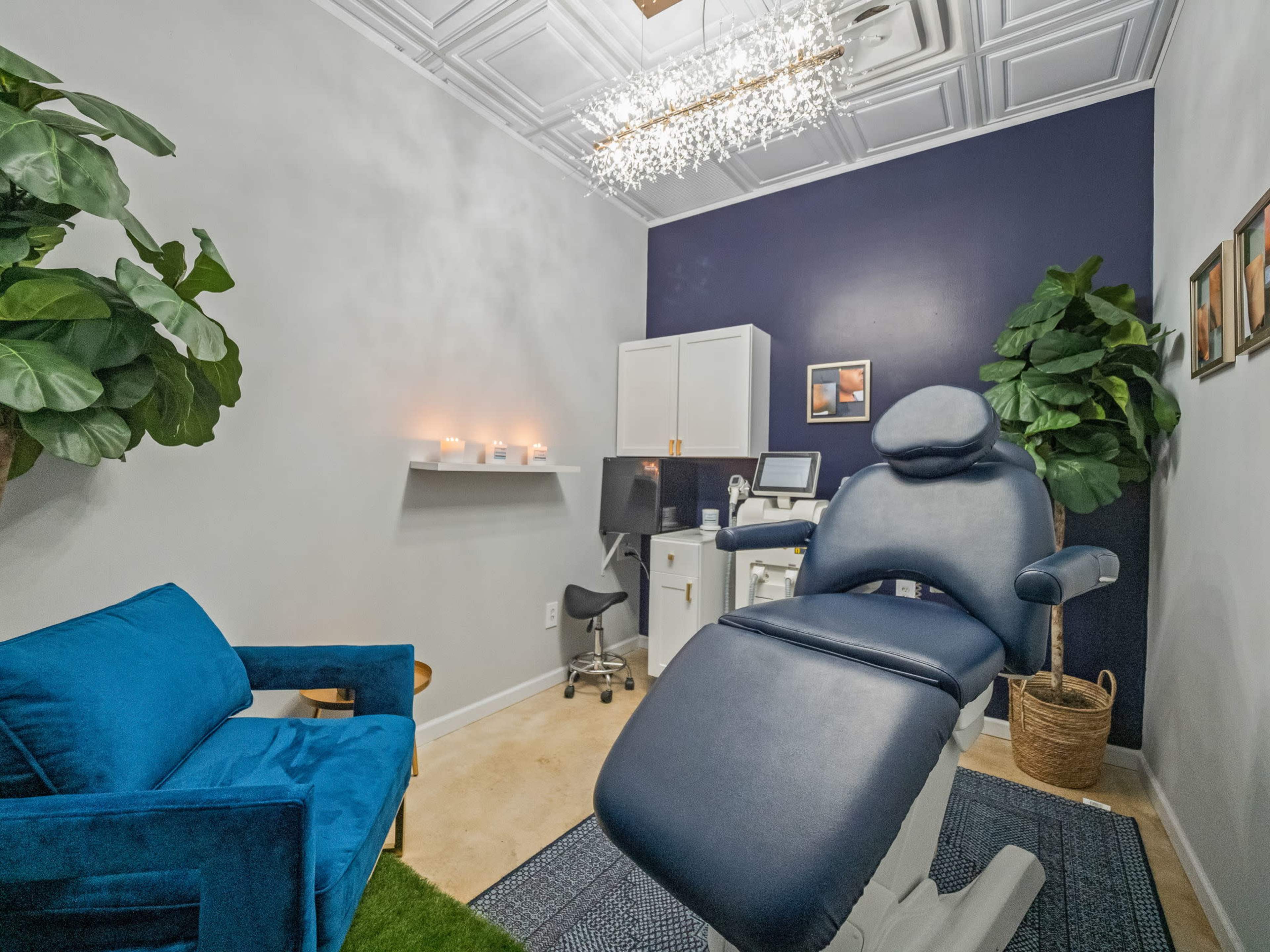 A small, well-decorated treatment room with a reclining chair, a desk with a computer, and decorative plants.
