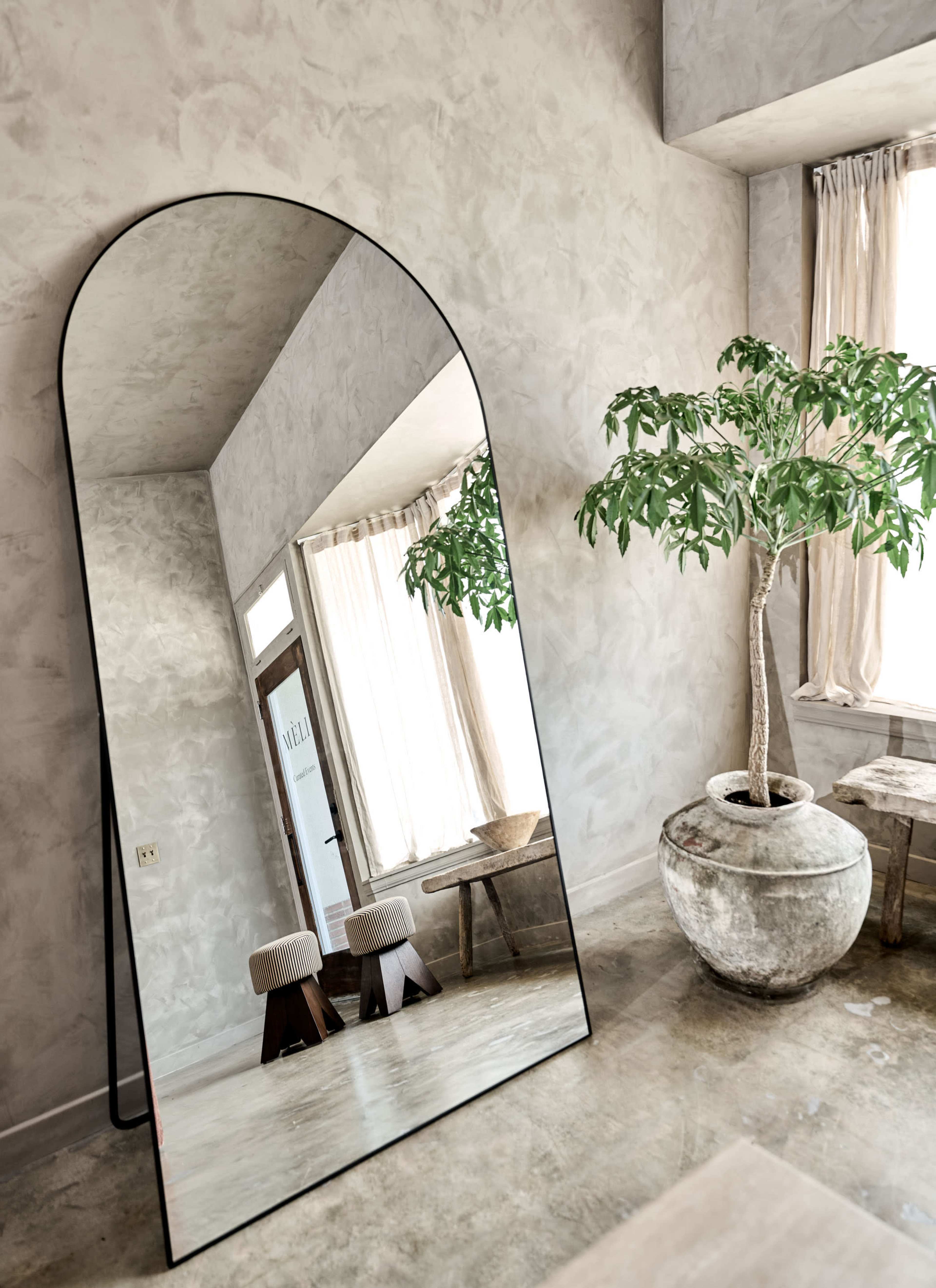 A large, arched mirror reflects a room with a potted plant, wooden stools, and natural light filtering through sheer curtains.