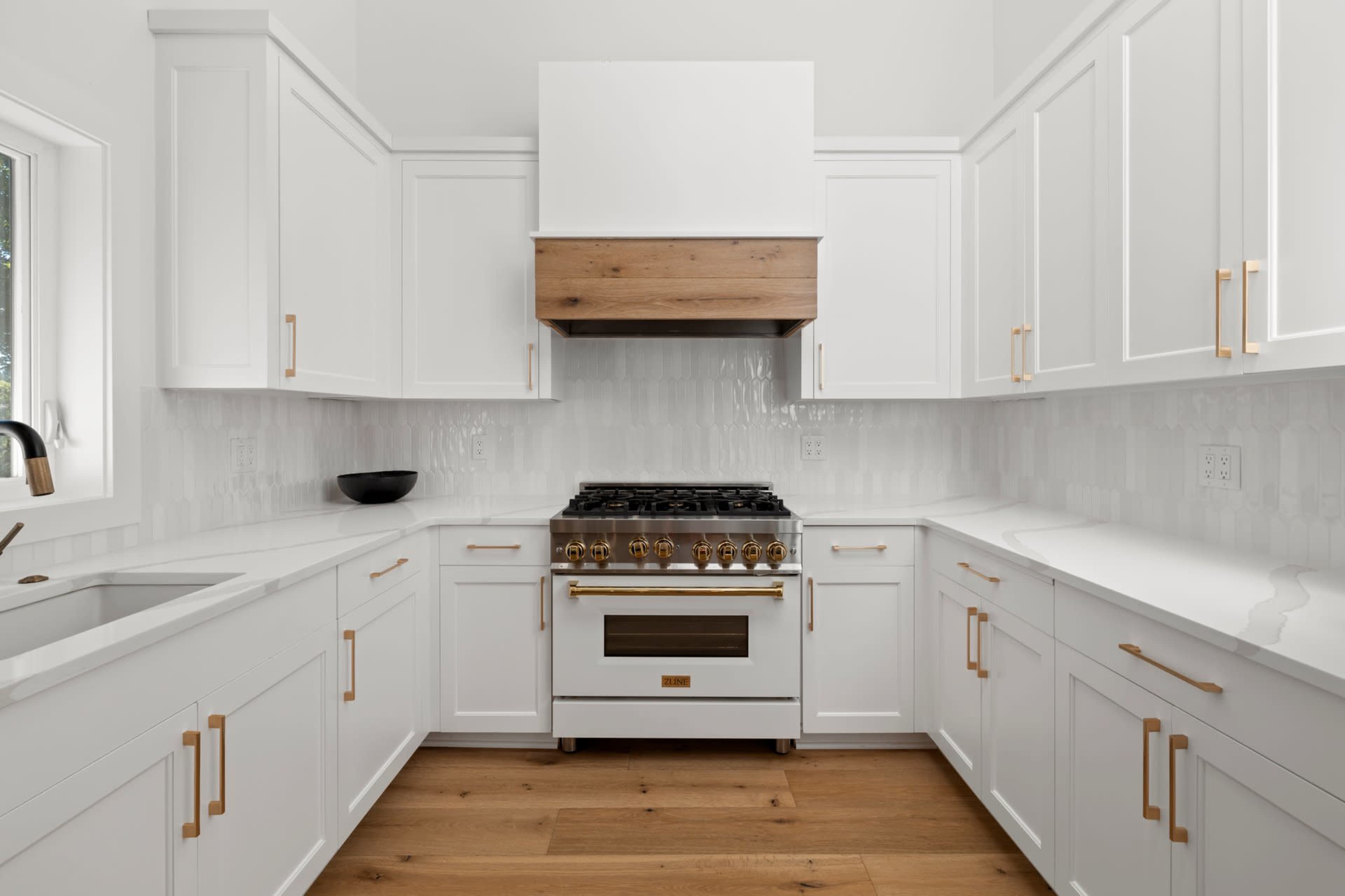 A modern kitchen features white cabinets, a wooden range hood, and a stainless steel stove.