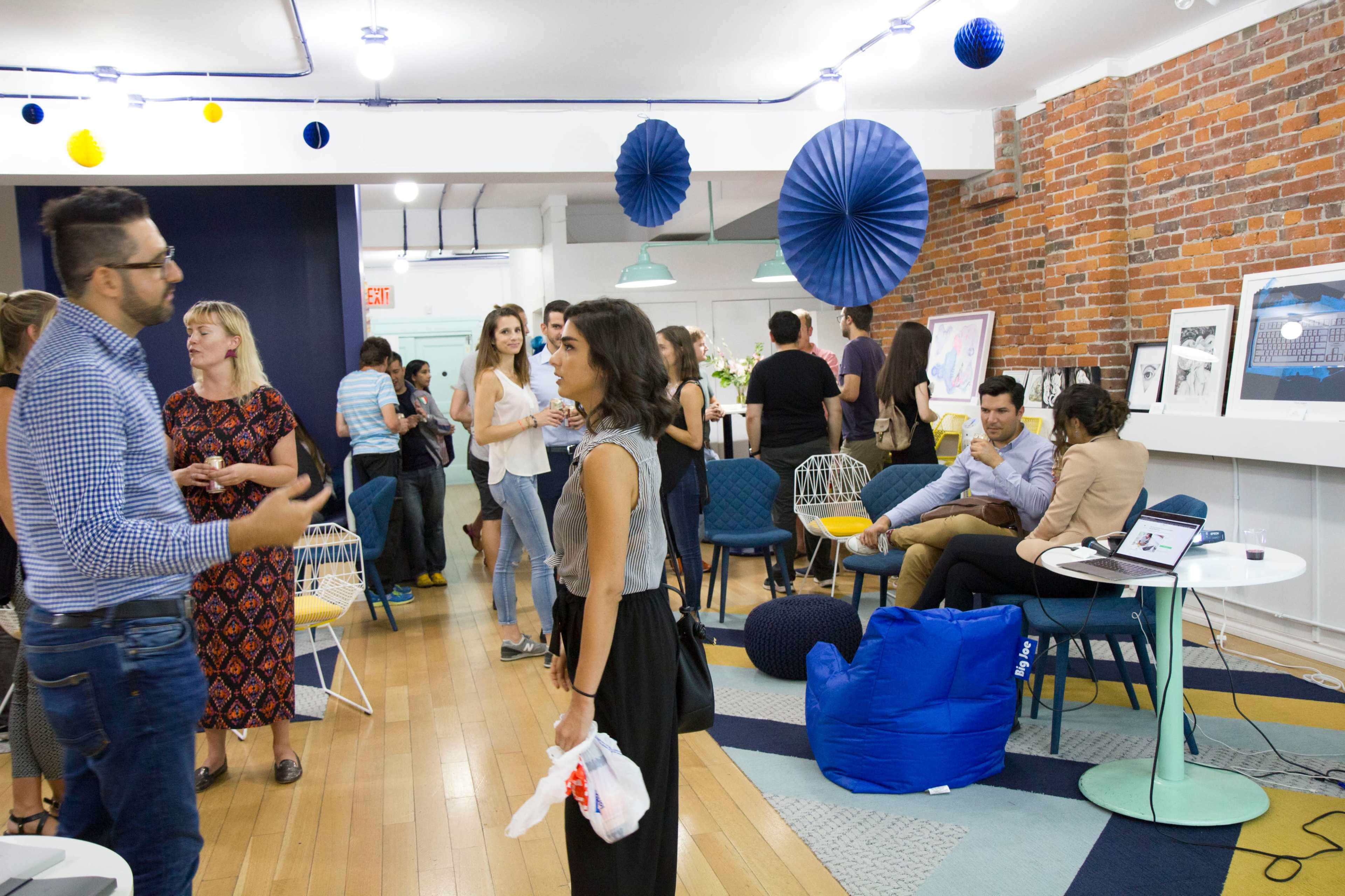 A group of people converses and socializes in a modern, decorated indoor space with blue and yellow decorations and a mix of seating arrangements.