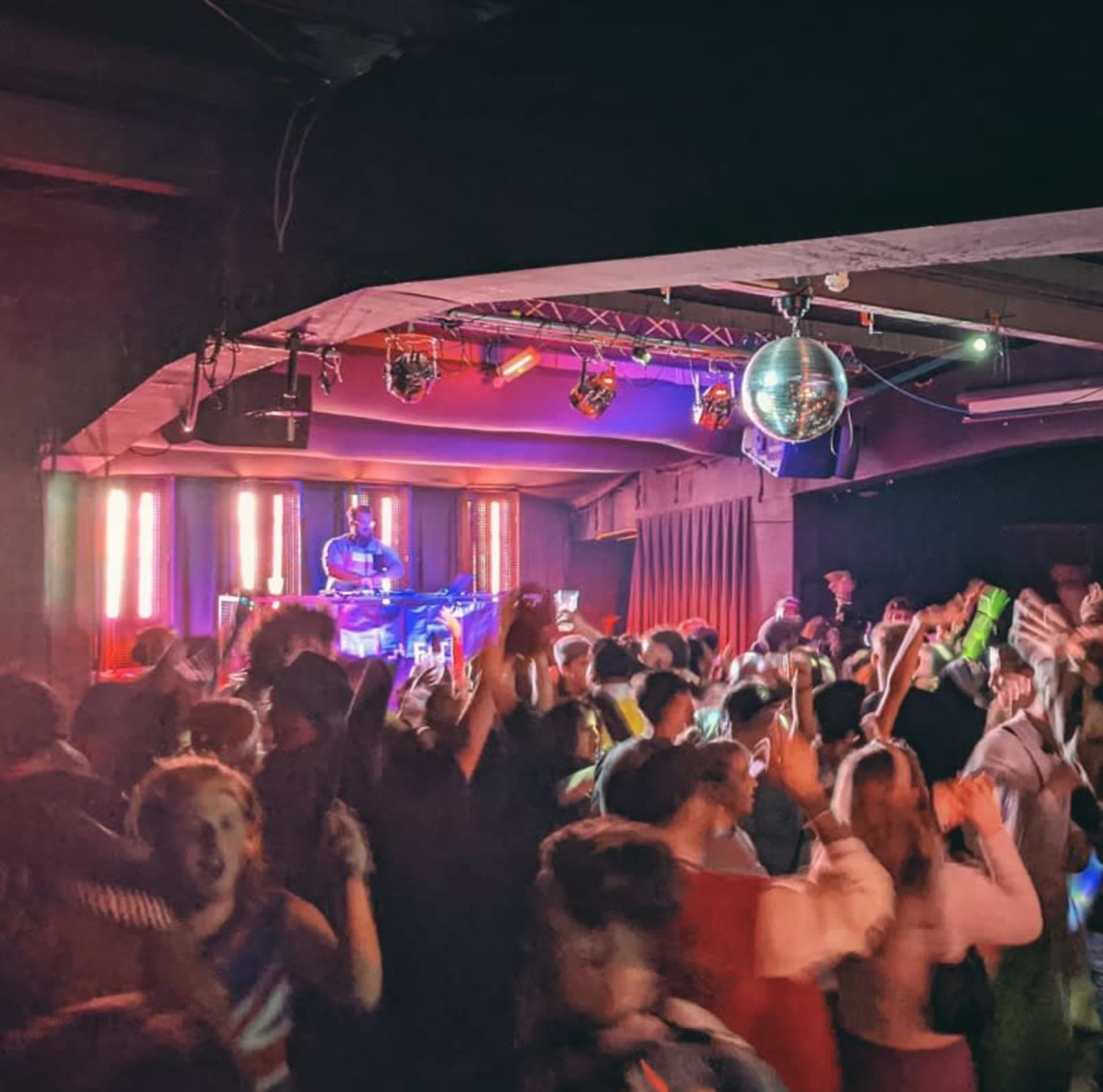 A crowded nightclub scene features people dancing energetically while a DJ performs on stage, illuminated by colorful lights and a disco ball overhead.
