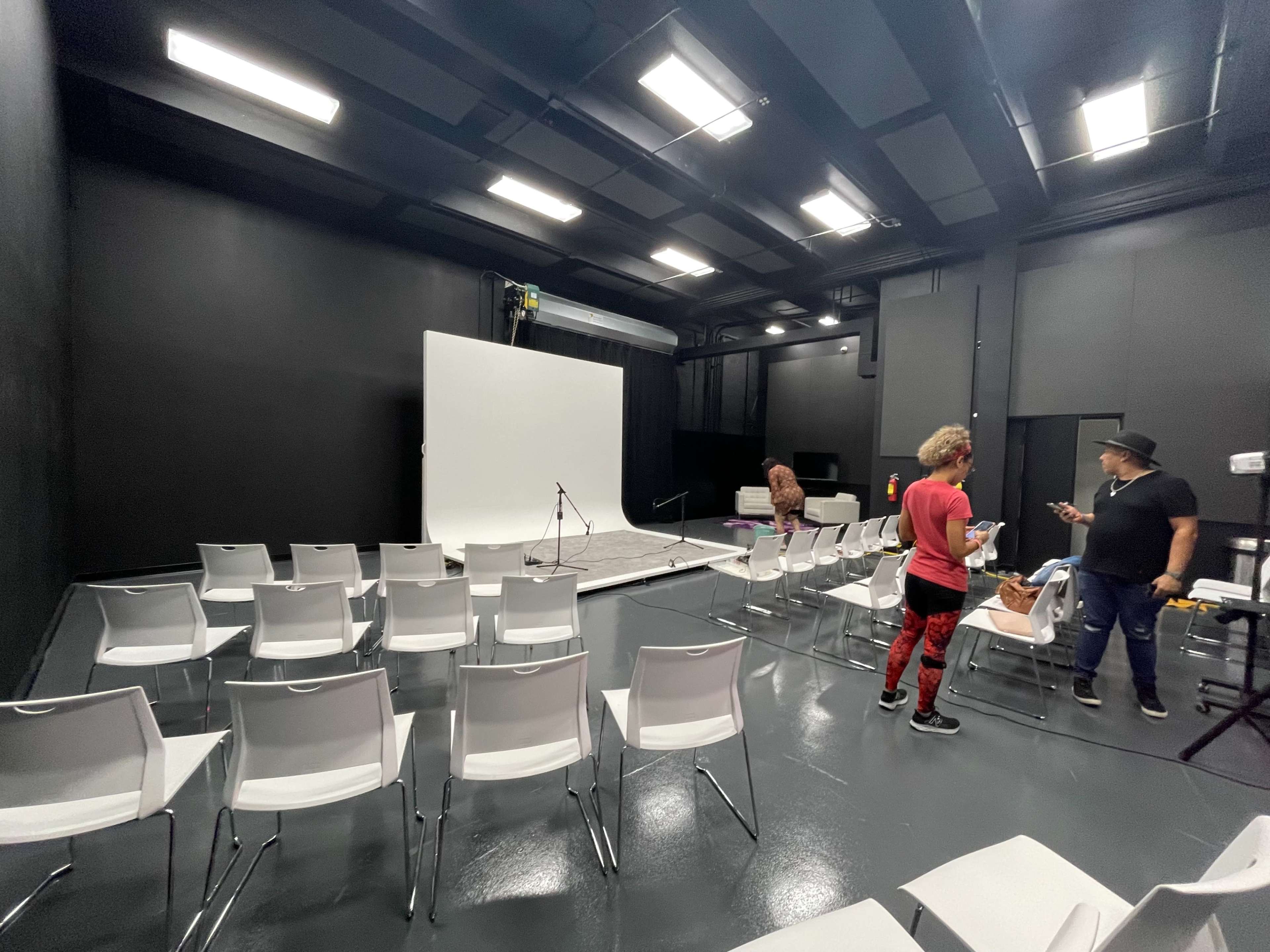 A spacious studio features white chairs arranged in rows facing a blank backdrop, with two individuals engaged in conversation near a setup area.