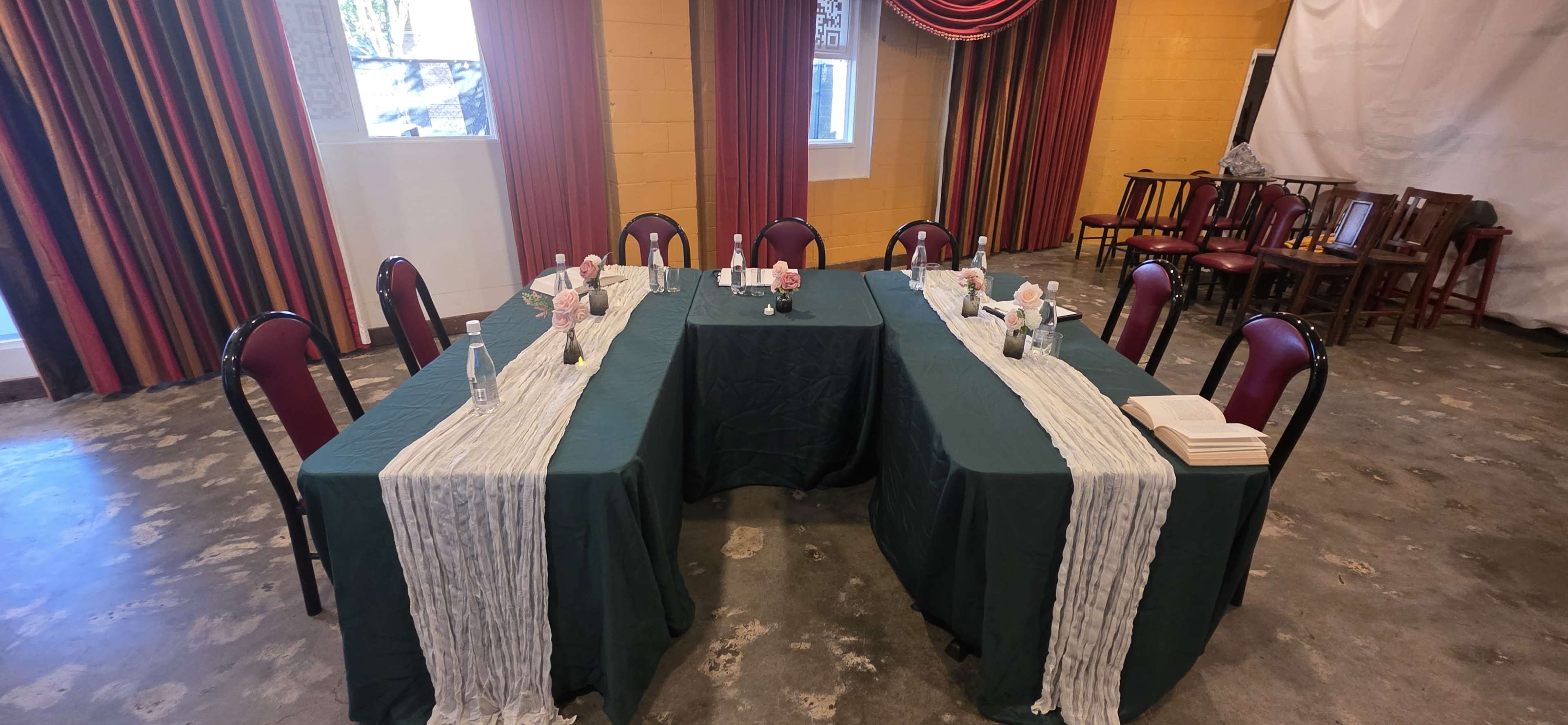 Two rectangular tables covered with green tablecloths are arranged in a squared formation, adorned with floral centerpieces and flanked by maroon chairs in a room with golden-yellow walls and patterned curtains.