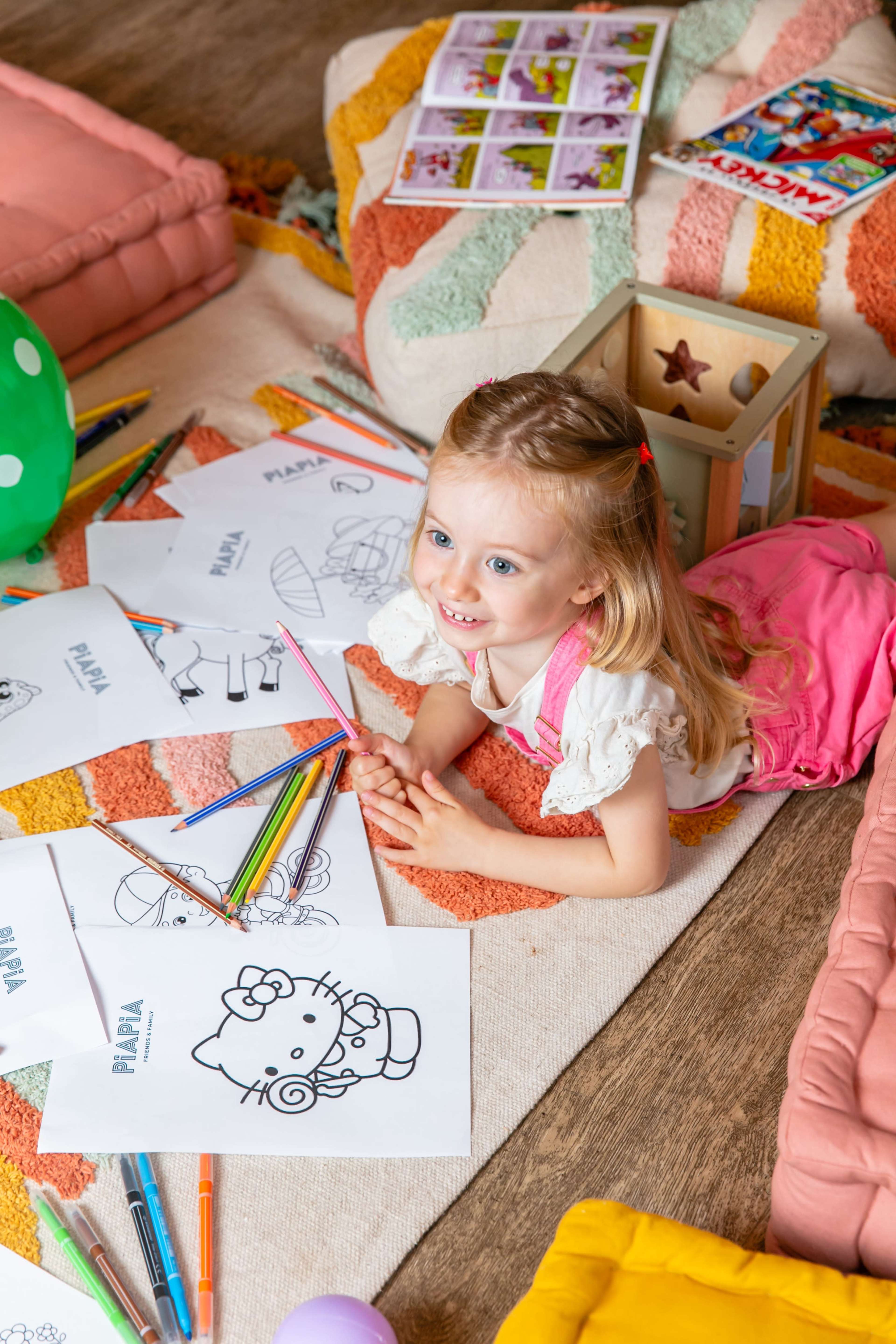 A young girl lies on a colorful rug surrounded by coloring sheets and art supplies.