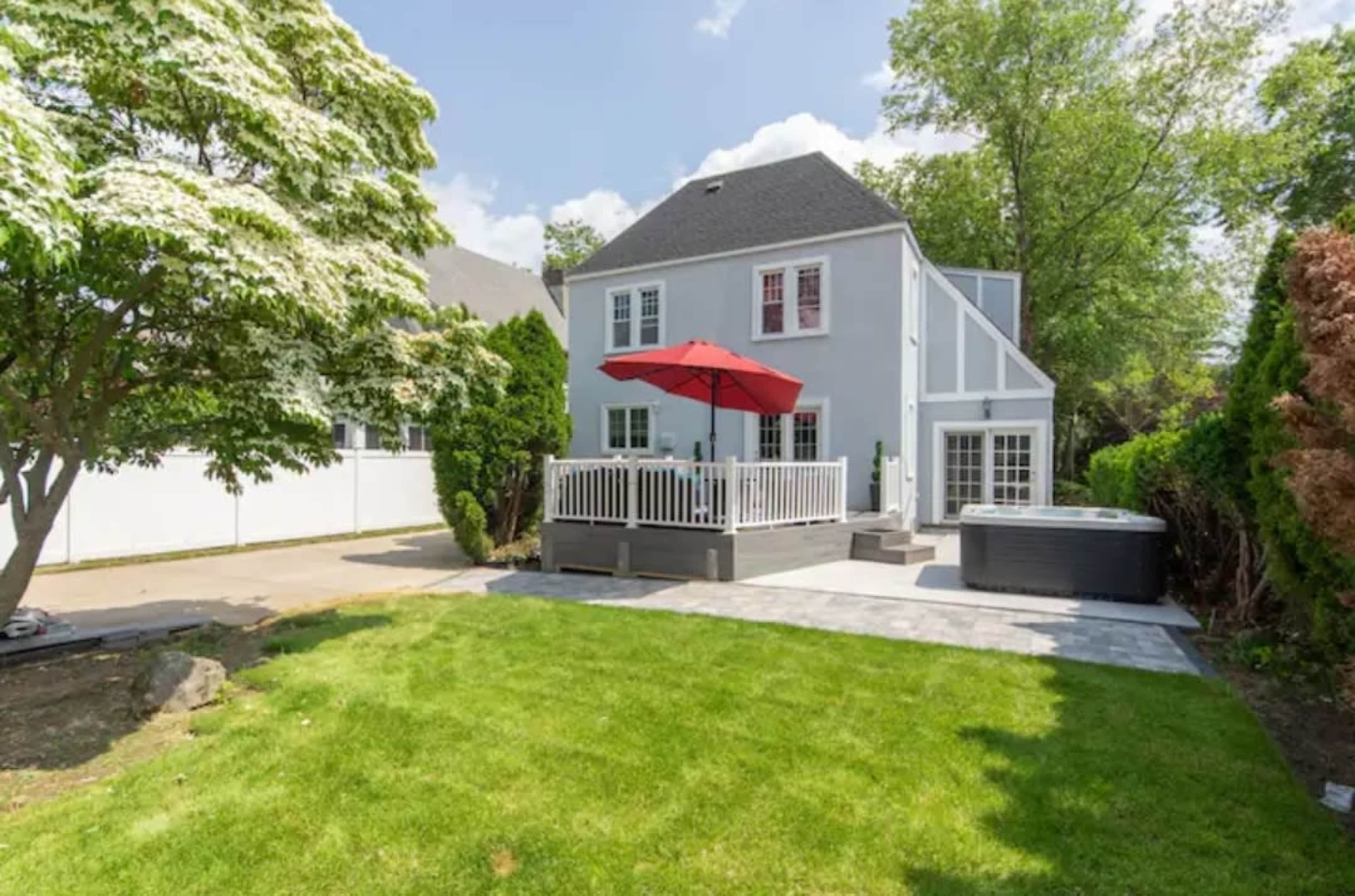 A house with a gray exterior, a deck featuring a red umbrella, and a hot tub, all set in a well-maintained yard with green grass and trees.