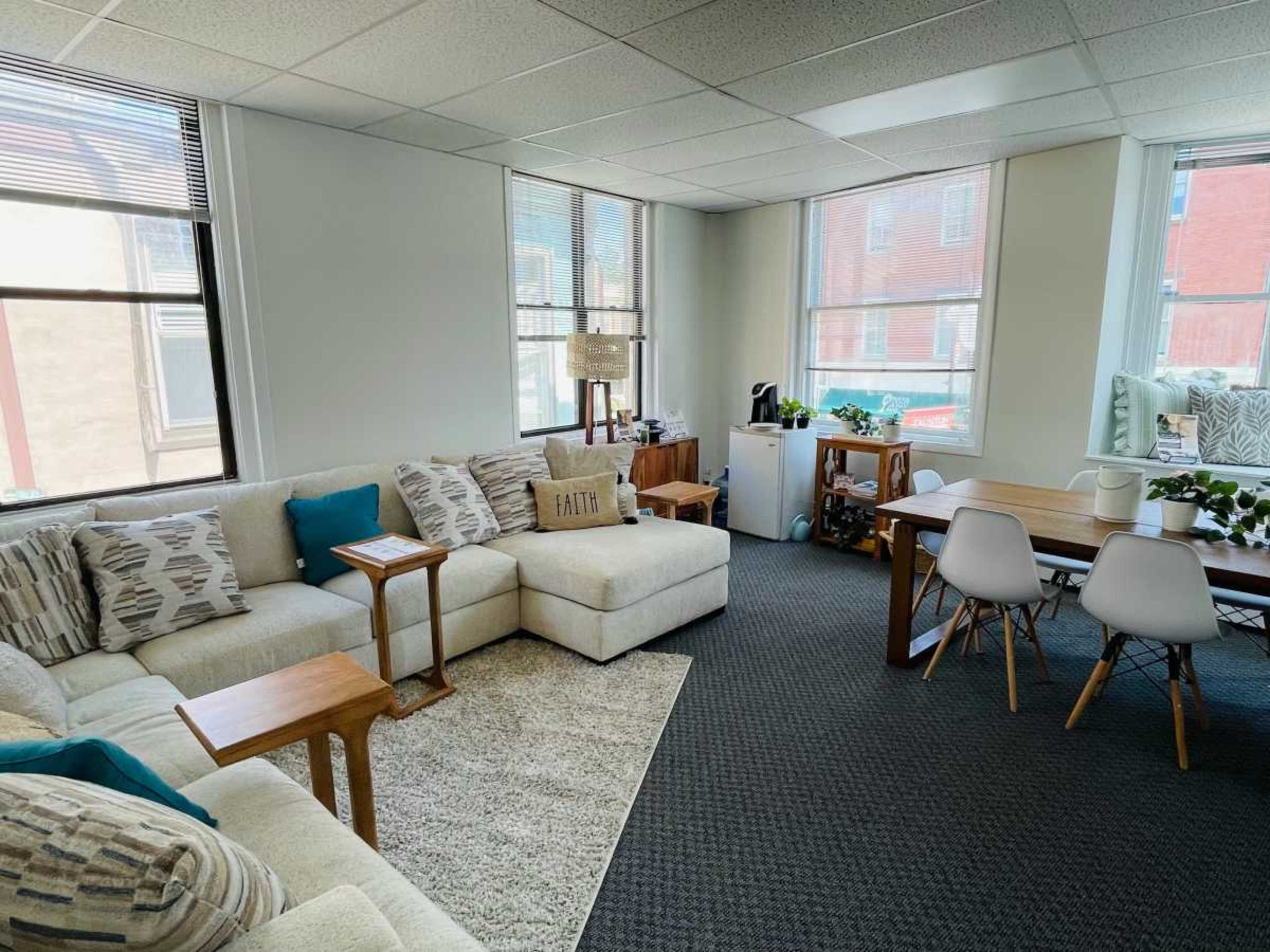 A bright, modern living space features a white sectional sofa, a small dining table, and large windows that let in natural light.