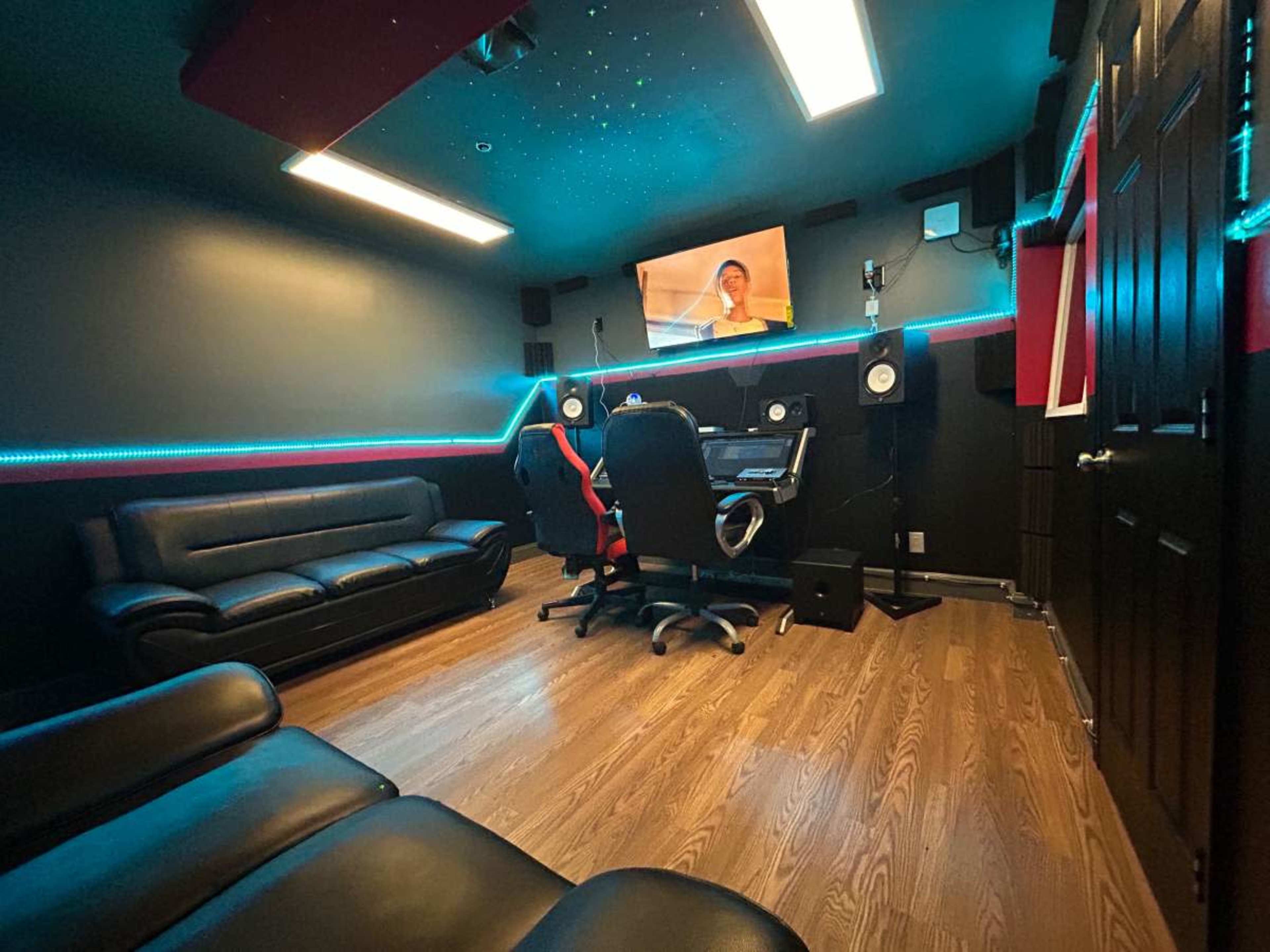 The image shows a small recording studio with black walls, modern furniture, a large screen displaying a video, and colorful LED lighting.