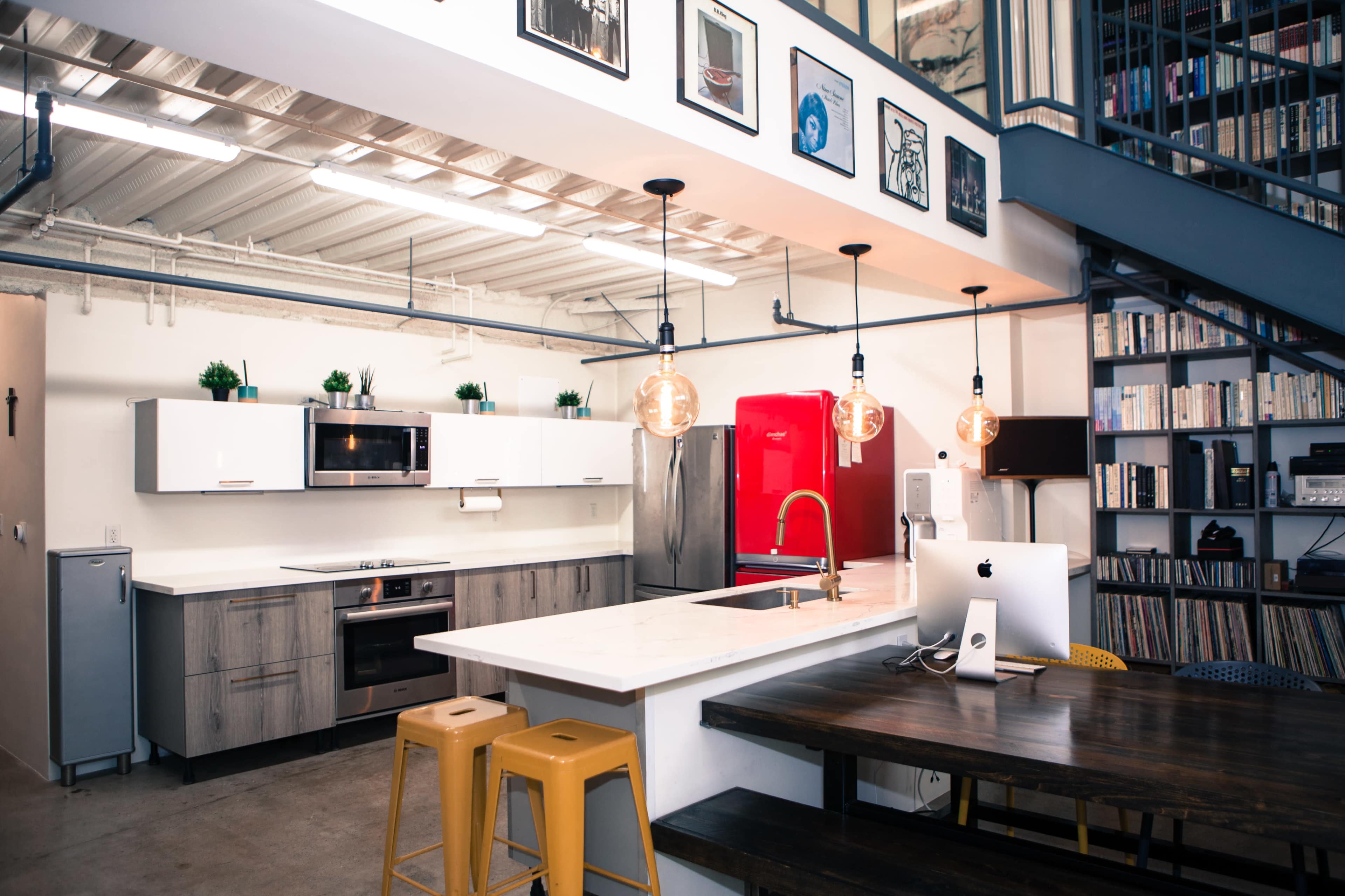 A modern kitchen with white cabinets, a red refrigerator, a white countertop, and a wooden dining table under hanging light fixtures.