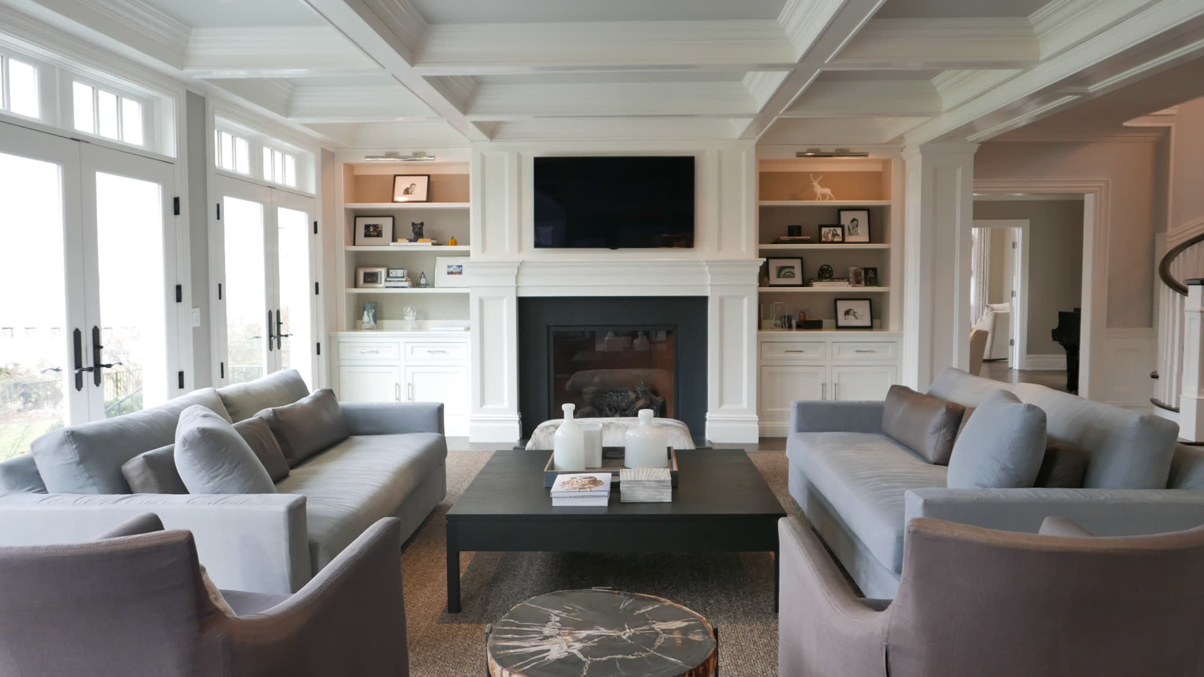 The image shows a modern living room with two gray sofas facing a coffee table, a fireplace, and built-in shelves displaying various items.