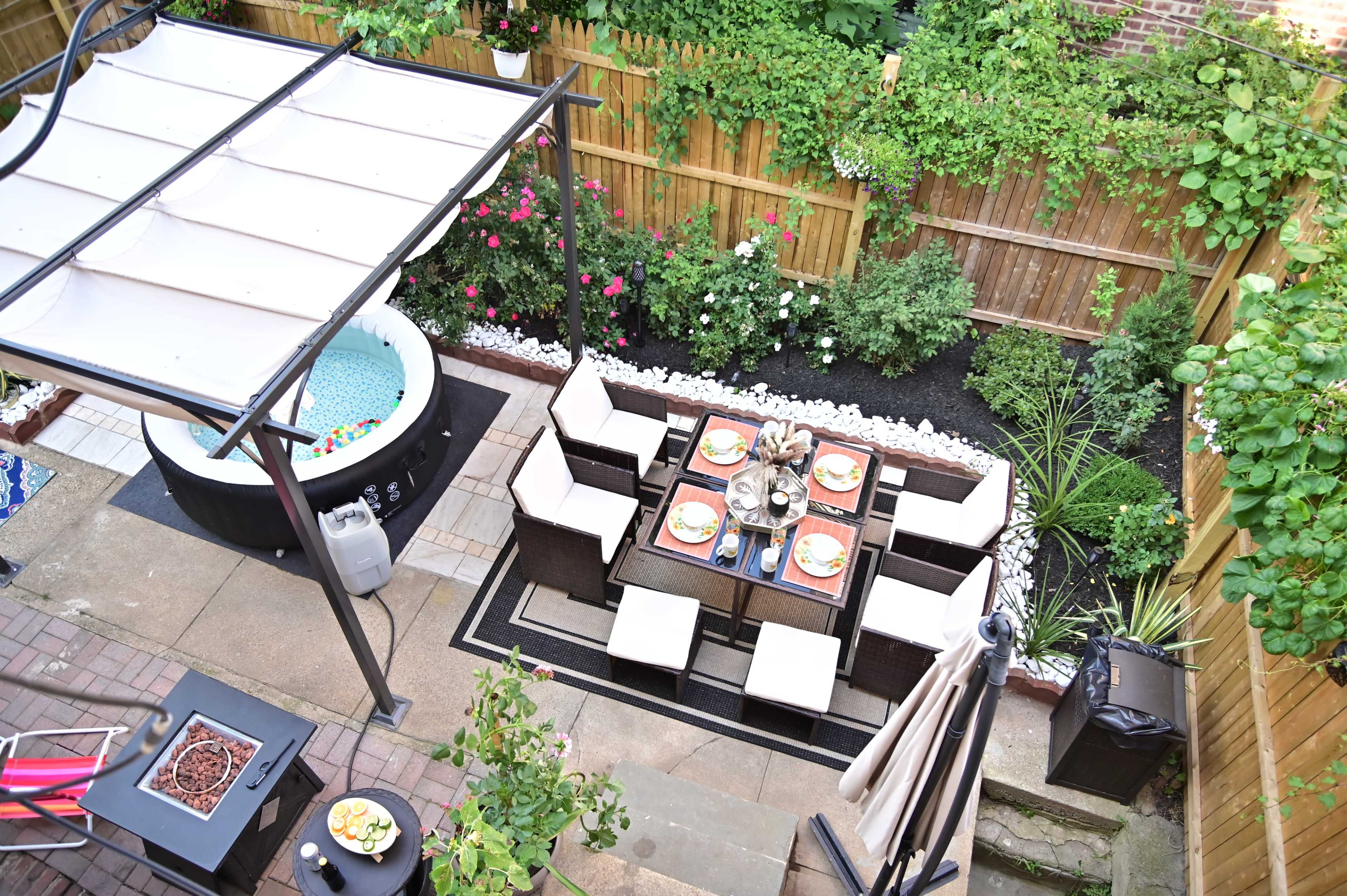 The image shows a neatly arranged outdoor patio with a dining set, a hot tub, and landscaped greenery surrounded by a wooden fence.