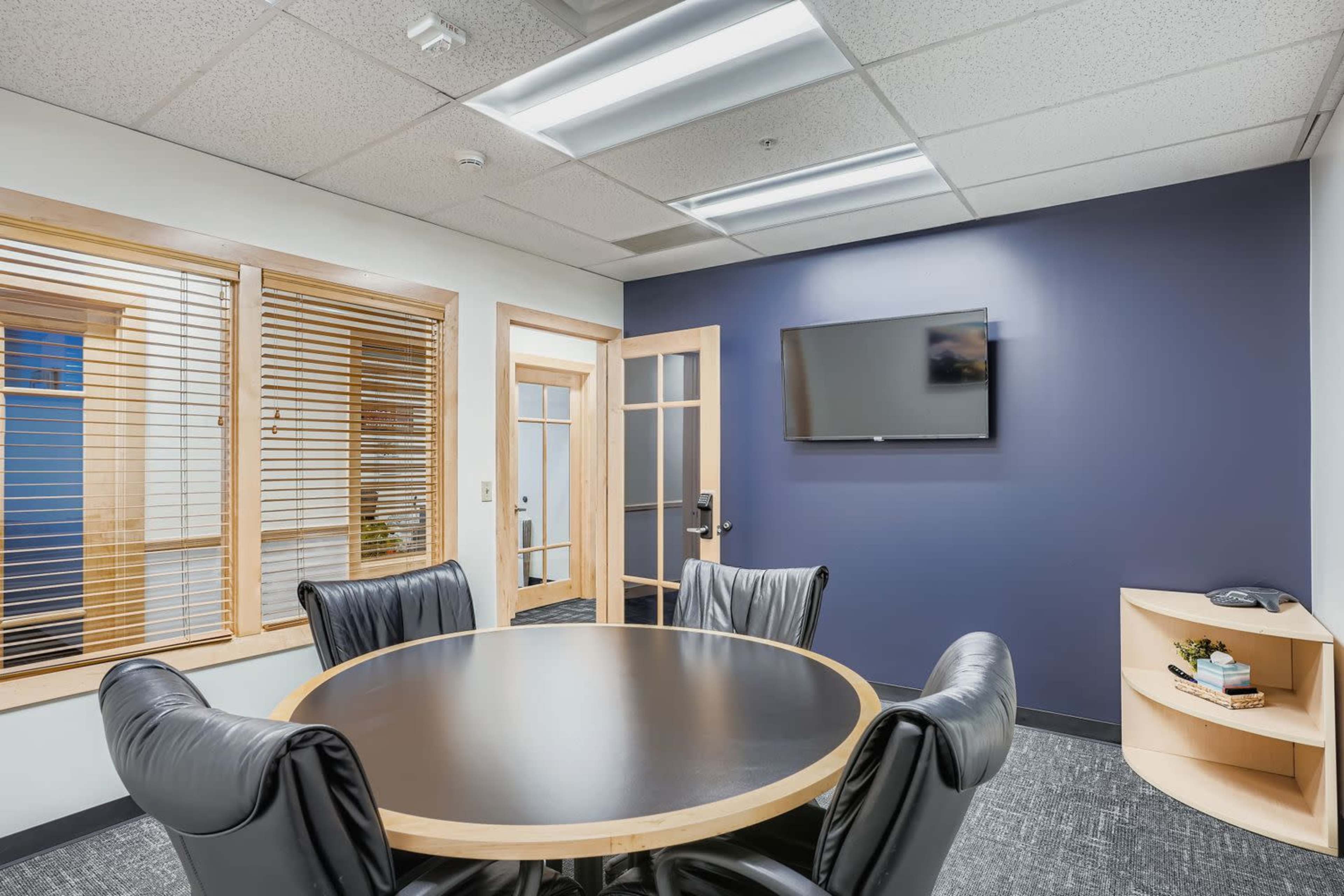 The image shows a conference room with a round table, black leather chairs, a television mounted on a dark blue wall, and large windows with blinds.