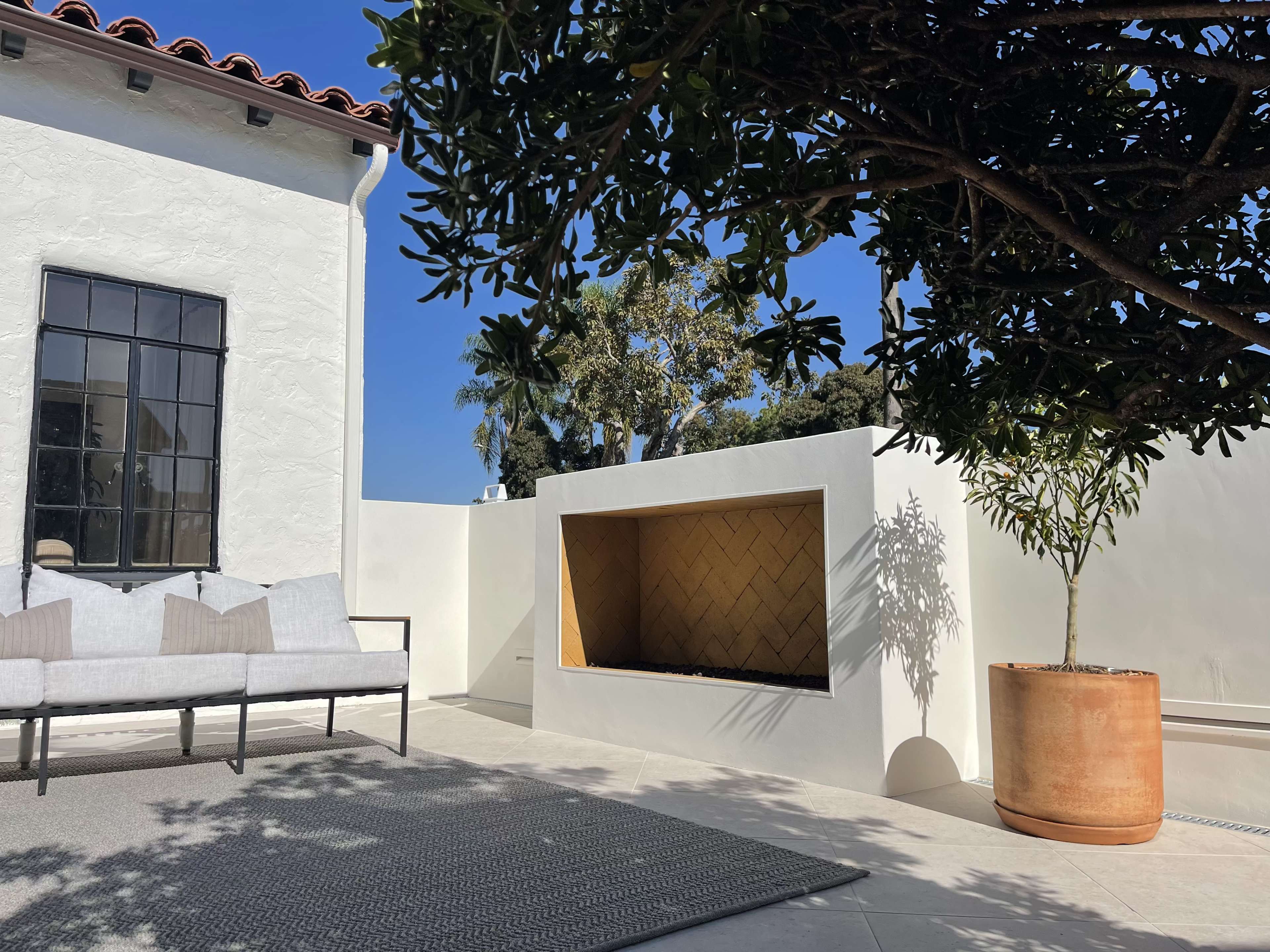 A modern outdoor space features a sleek, white wall with a recessed fireplace, a gray rug, a sofa, and a large potted plant under a tree.