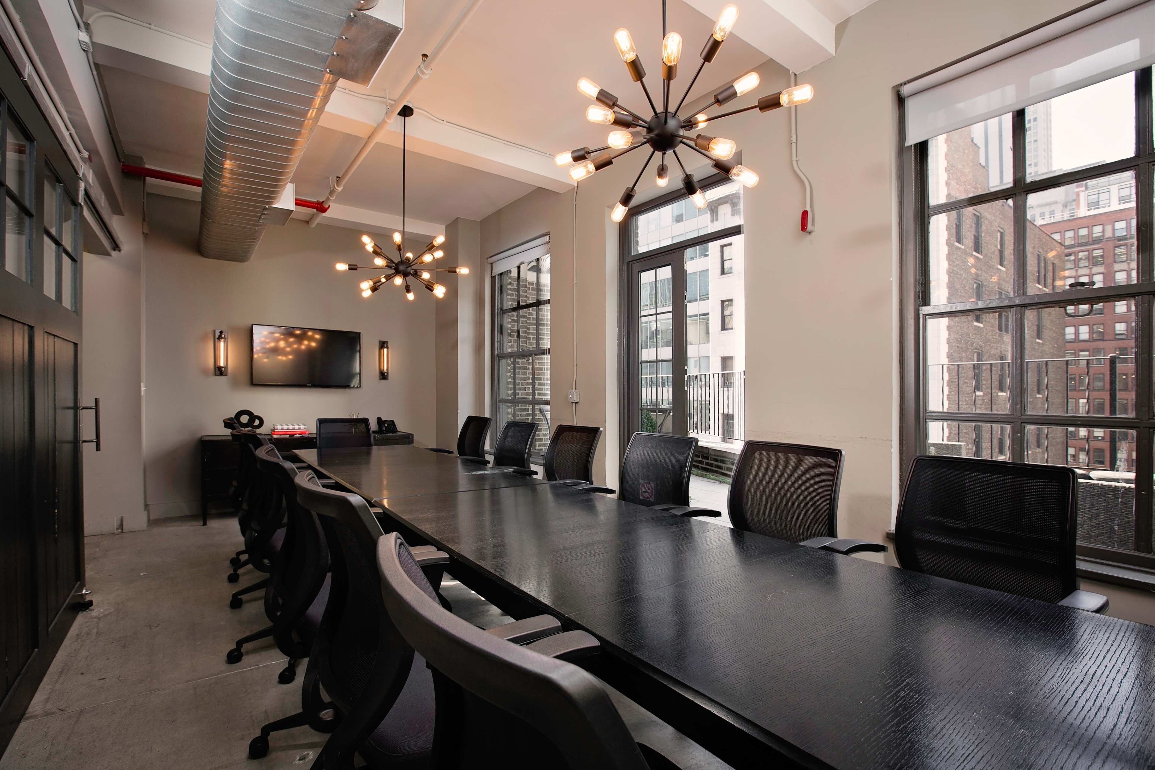 A modern conference room featuring a long black table, black ergonomic chairs, and two unique light fixtures, with large windows providing views of a cityscape.