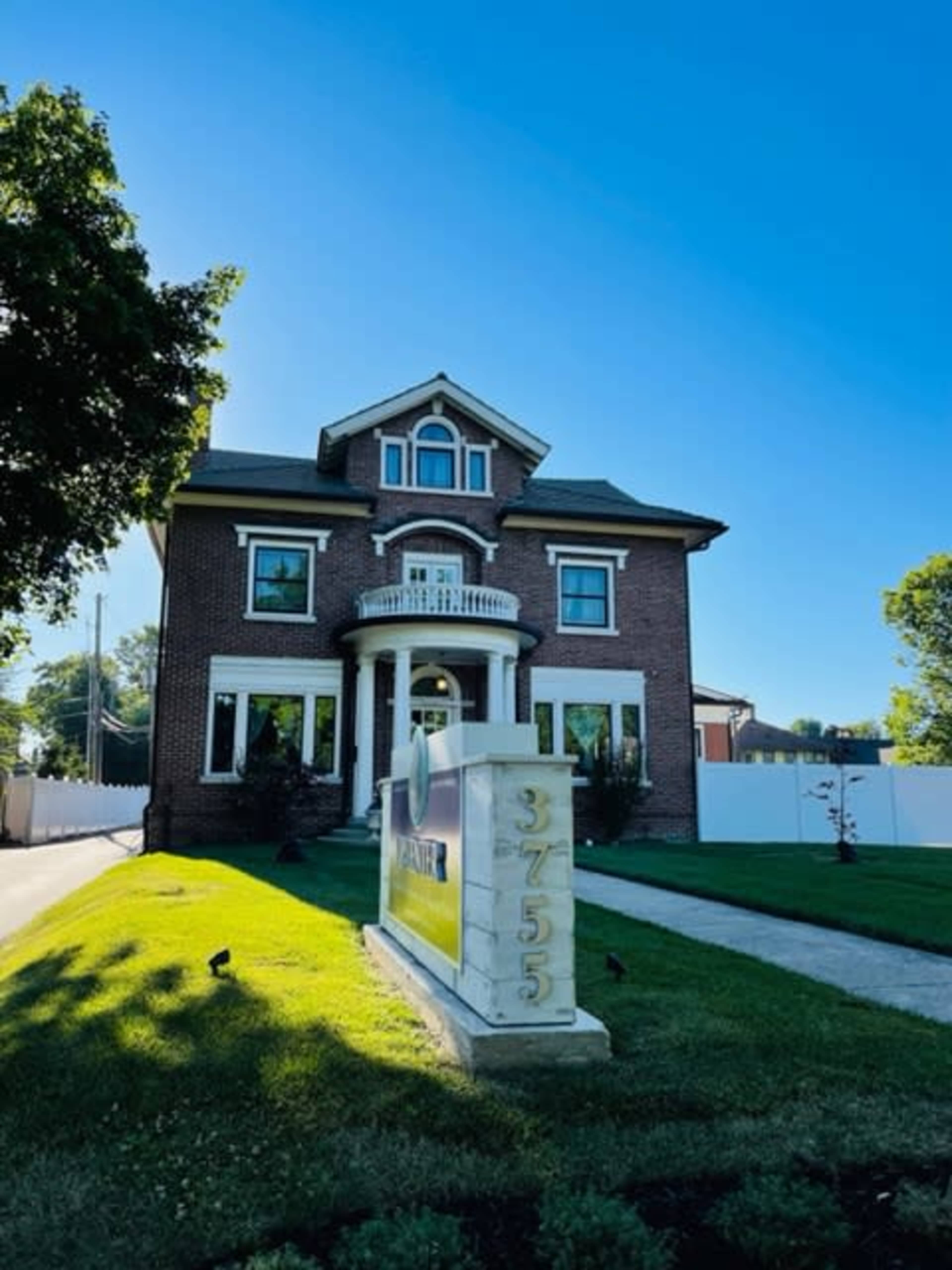 A large, two-story brick house with a front porch and well-maintained lawn stands beside a stone sign displaying the number 37555.