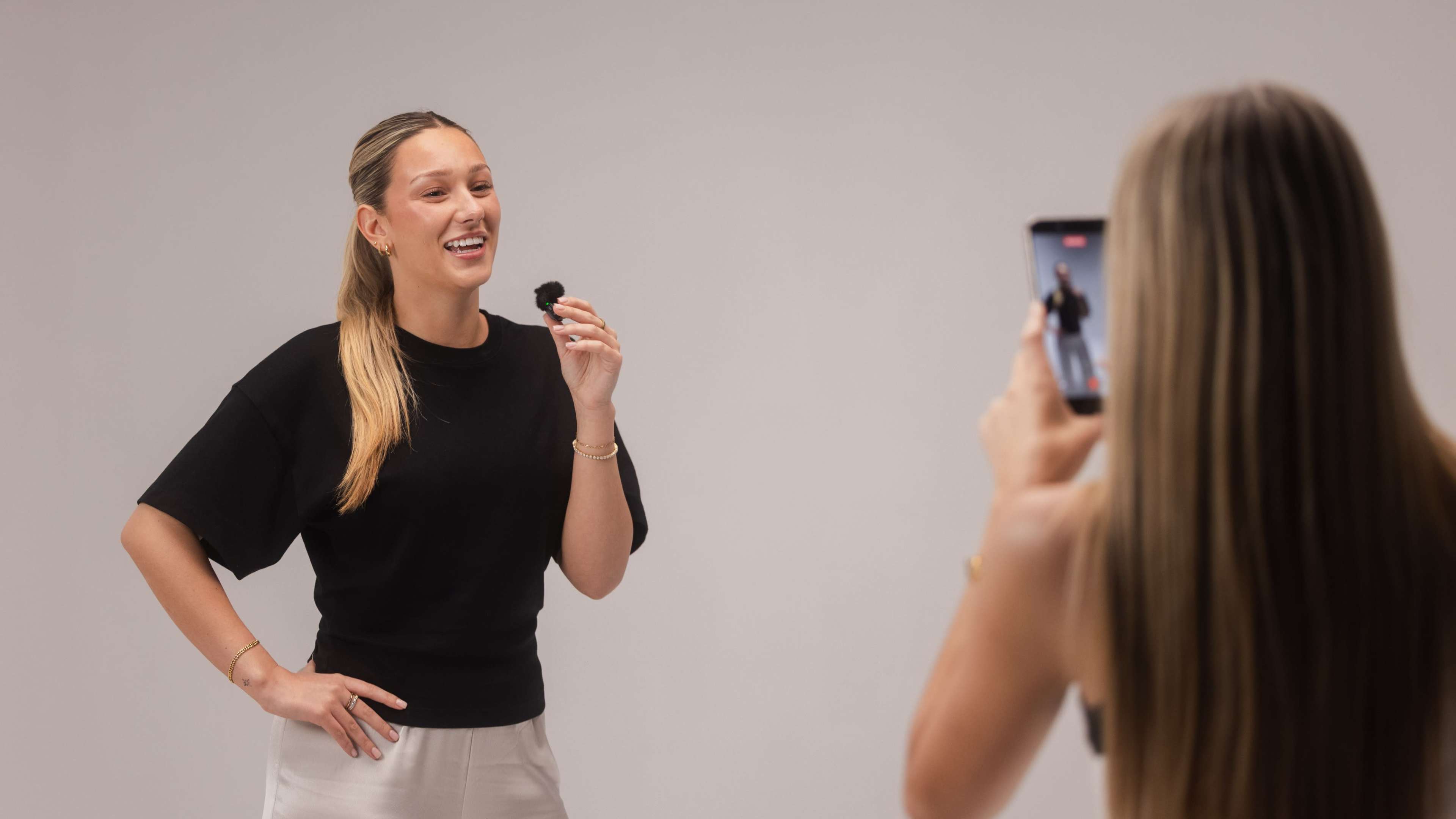 A woman holds a microphone while smiling and speaking, as another person takes a photo of her with a smartphone against a plain background.