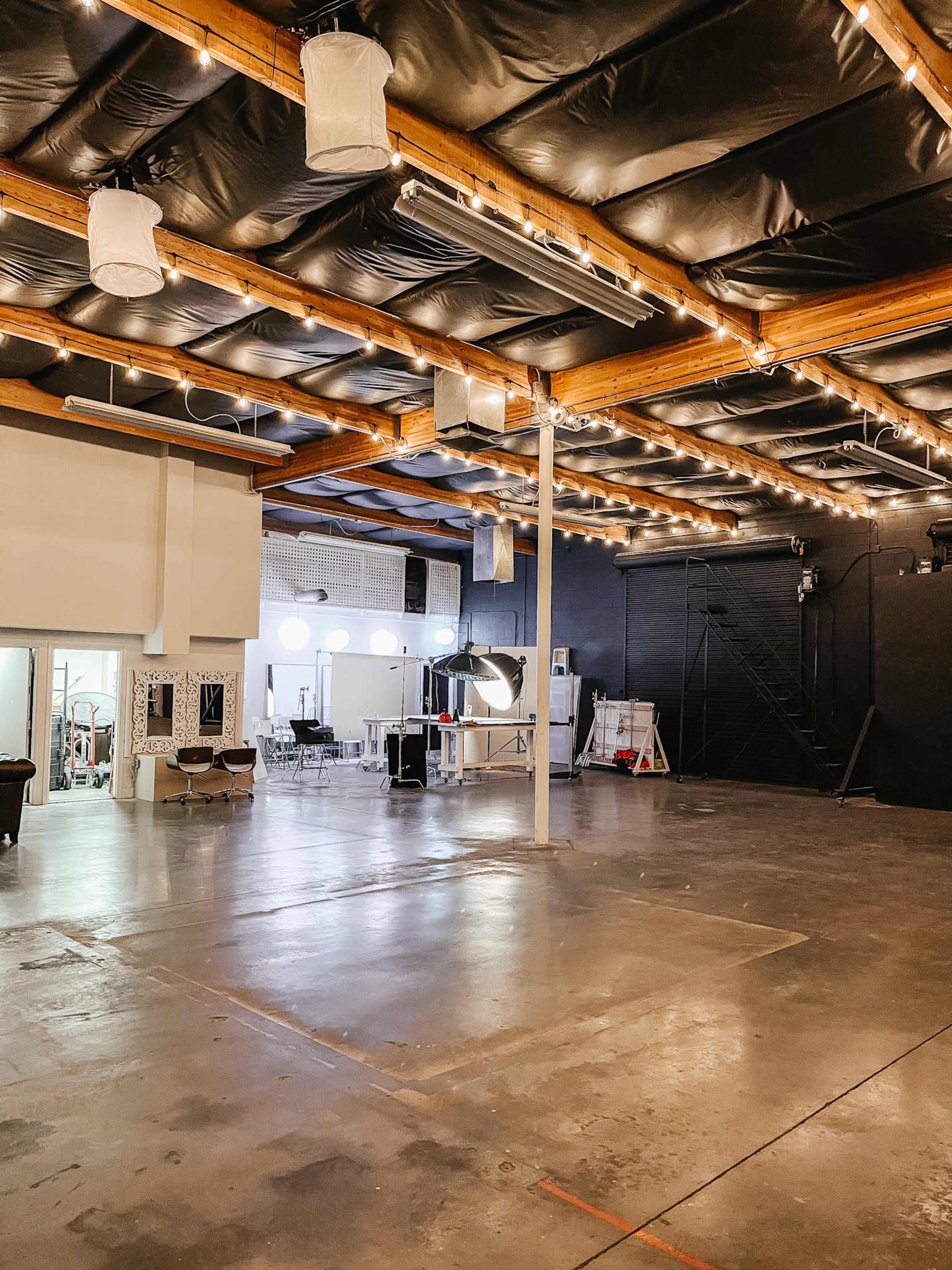 The image shows a spacious interior with a polished concrete floor, high ceilings, and string lights overhead, featuring a mix of studio equipment and furniture arranged in a creative workspace.
