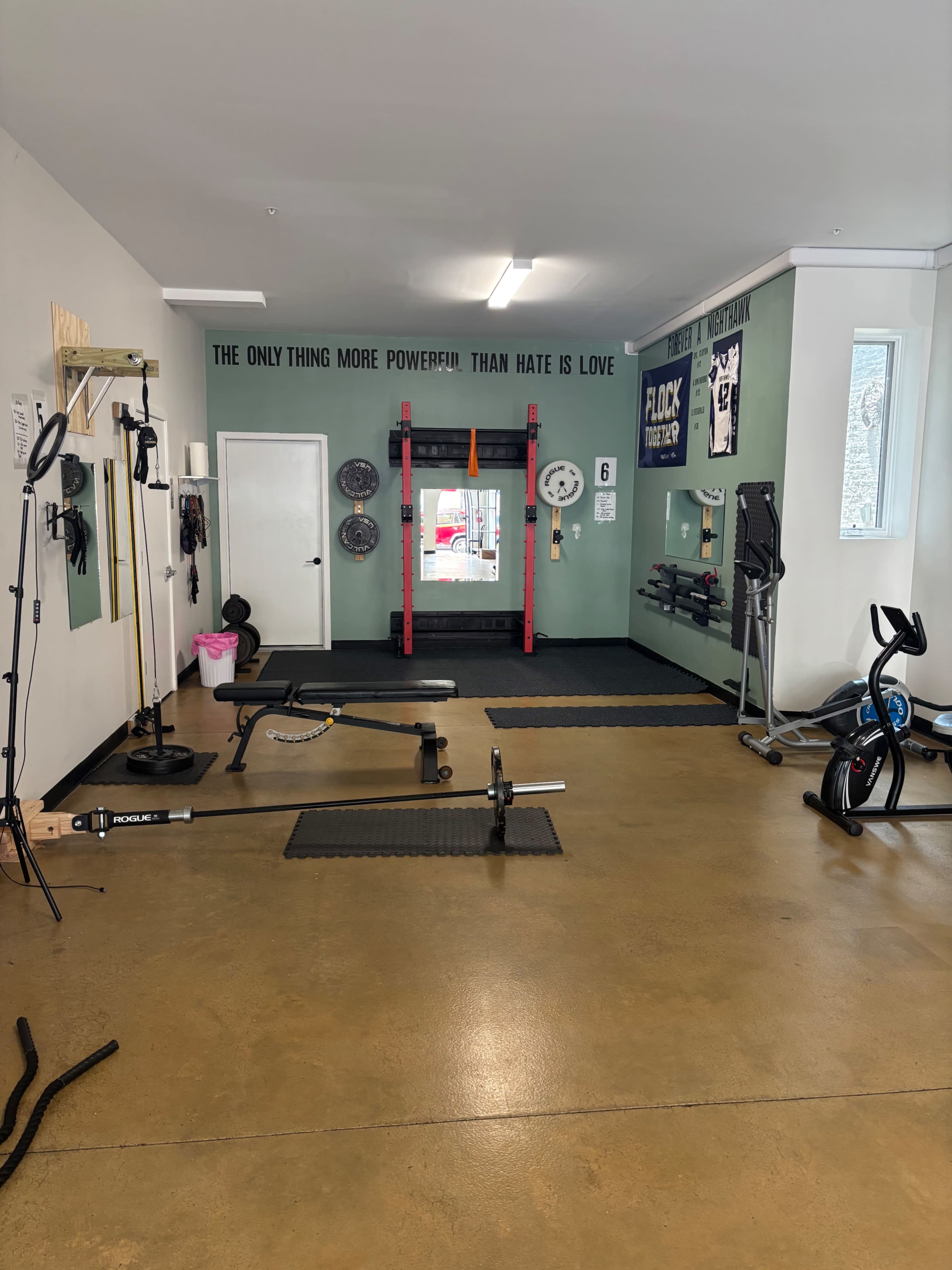 The image shows a functional gym space with weights, workout equipment, and a mirror, along with motivational text on the wall.