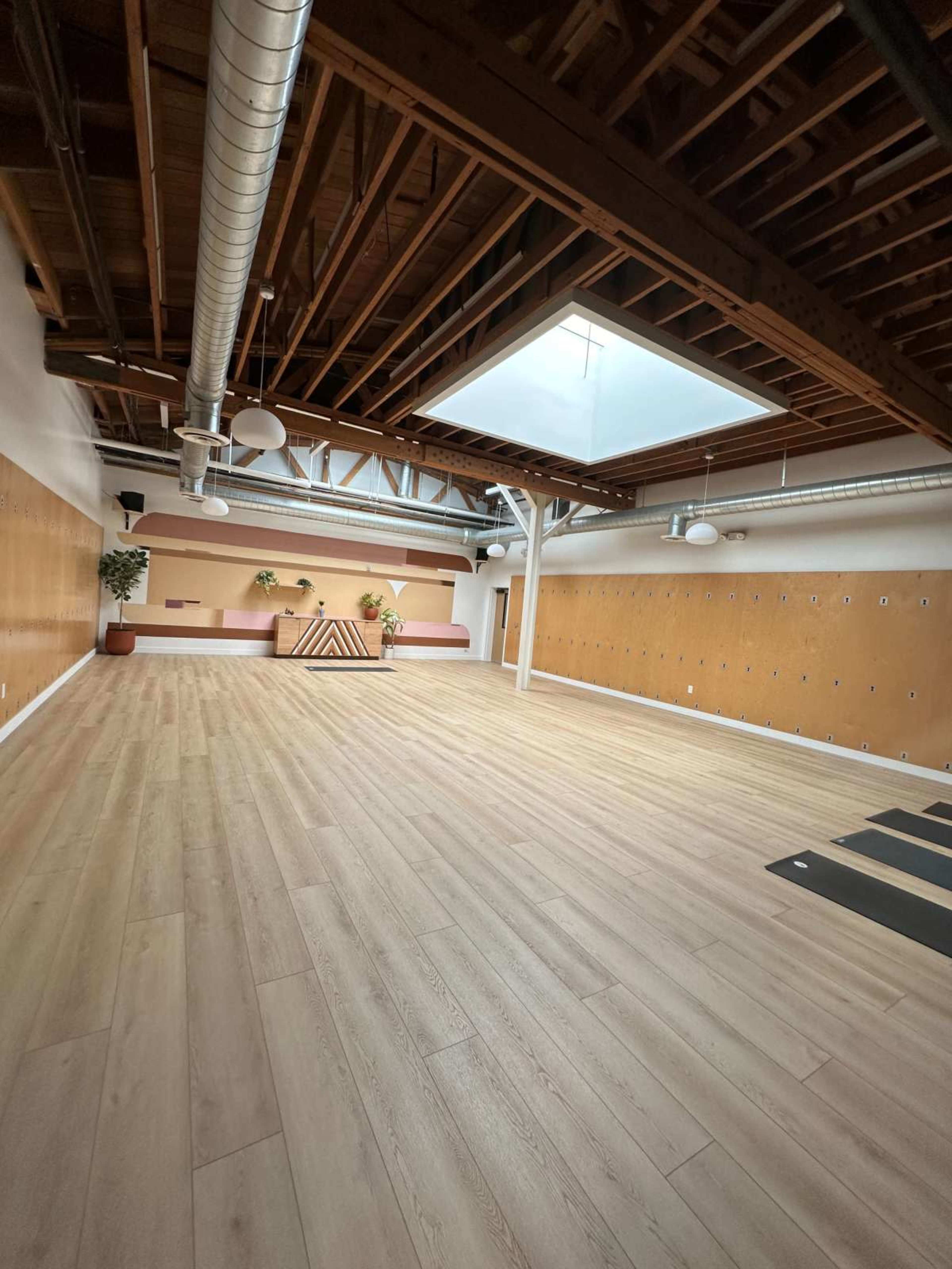 The image shows a spacious room with wooden flooring, a skylight, and minimalist decor, intended for yoga or fitness activities.