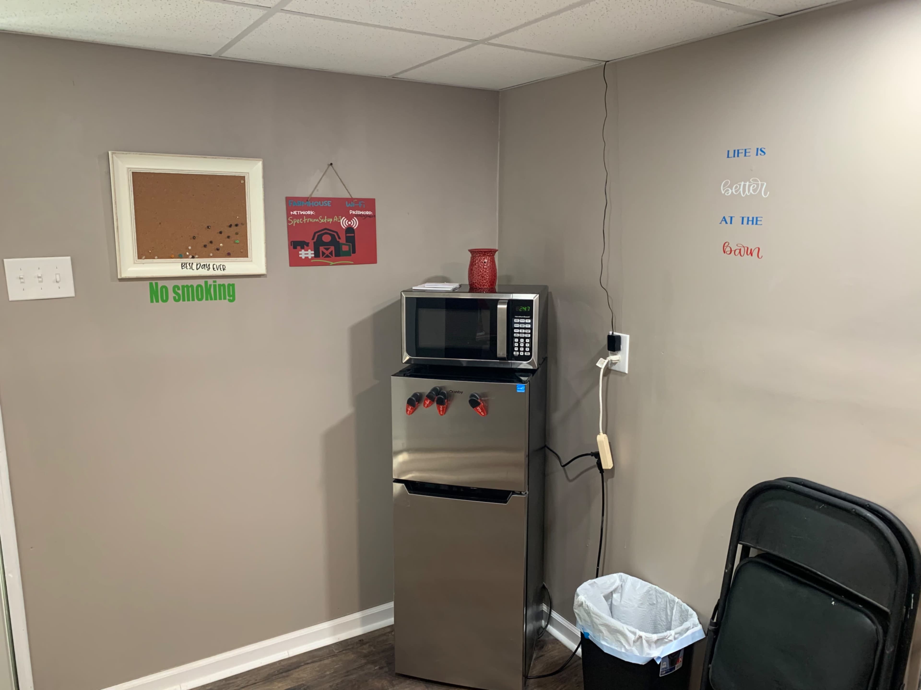 A small indoor area featuring a stainless steel refrigerator, a microwave on top, a corkboard on the wall, and a wastebasket beside a folding chair.