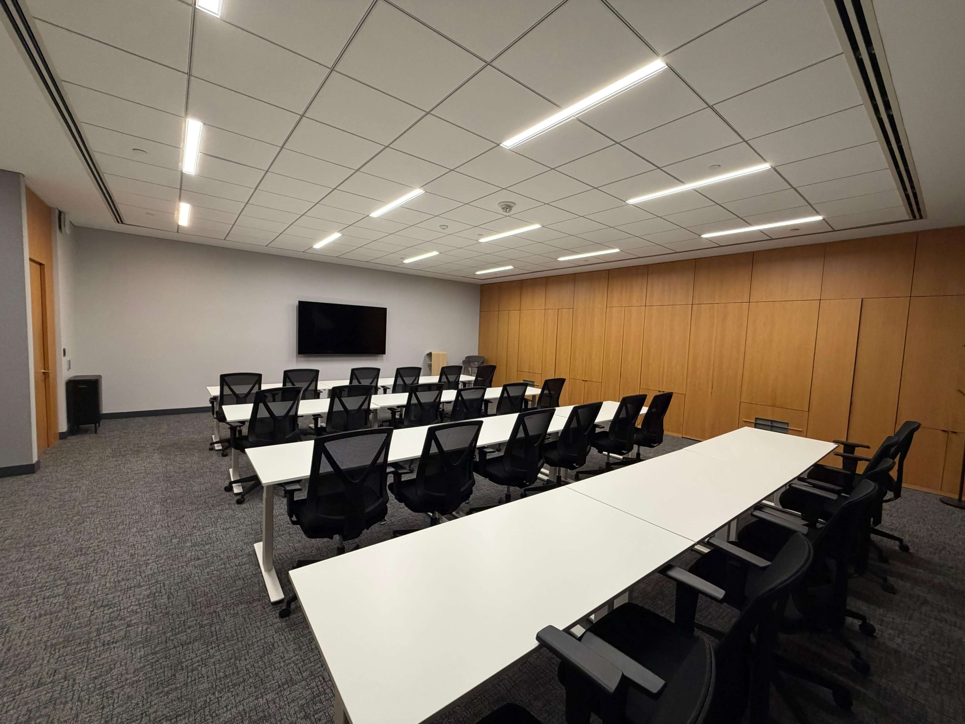 The image shows a modern conference room with a long, white table surrounded by black chairs, and a wall-mounted television on one side.